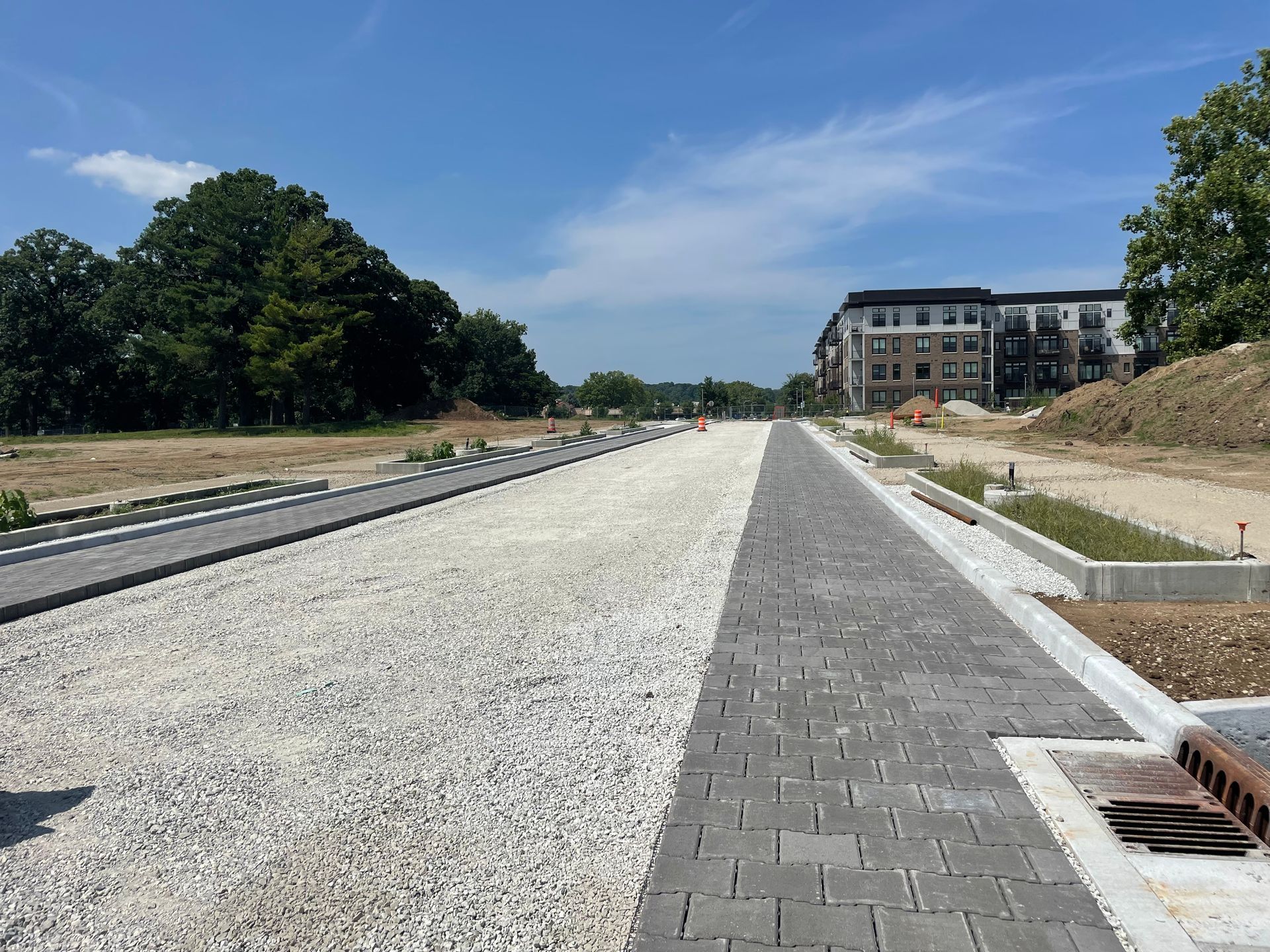 Wide gravel roadway under construction with paved edges, trees, and an apartment building in the distance