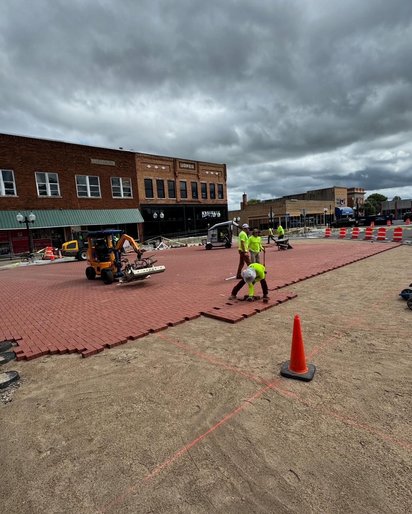 Workers paving a plaza with red bricks beside brick buildings under dark, cloudy skies.