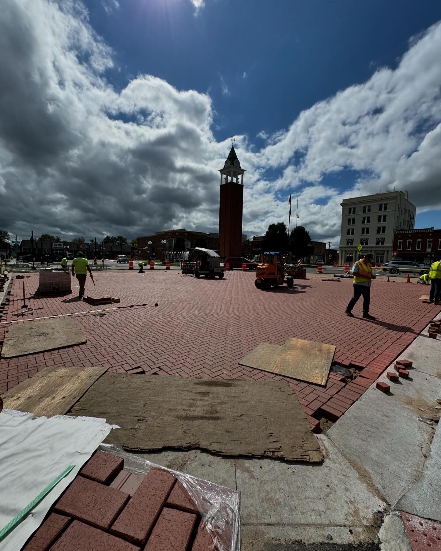 Town square with a tall clock tower, workers in safety vests, and red brick paving under a cloudy sky