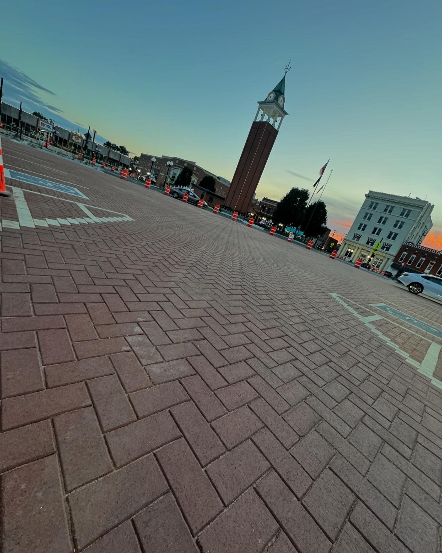 Tilted view of a brick plaza with a clock tower, buildings, and traffic cones at dusk
