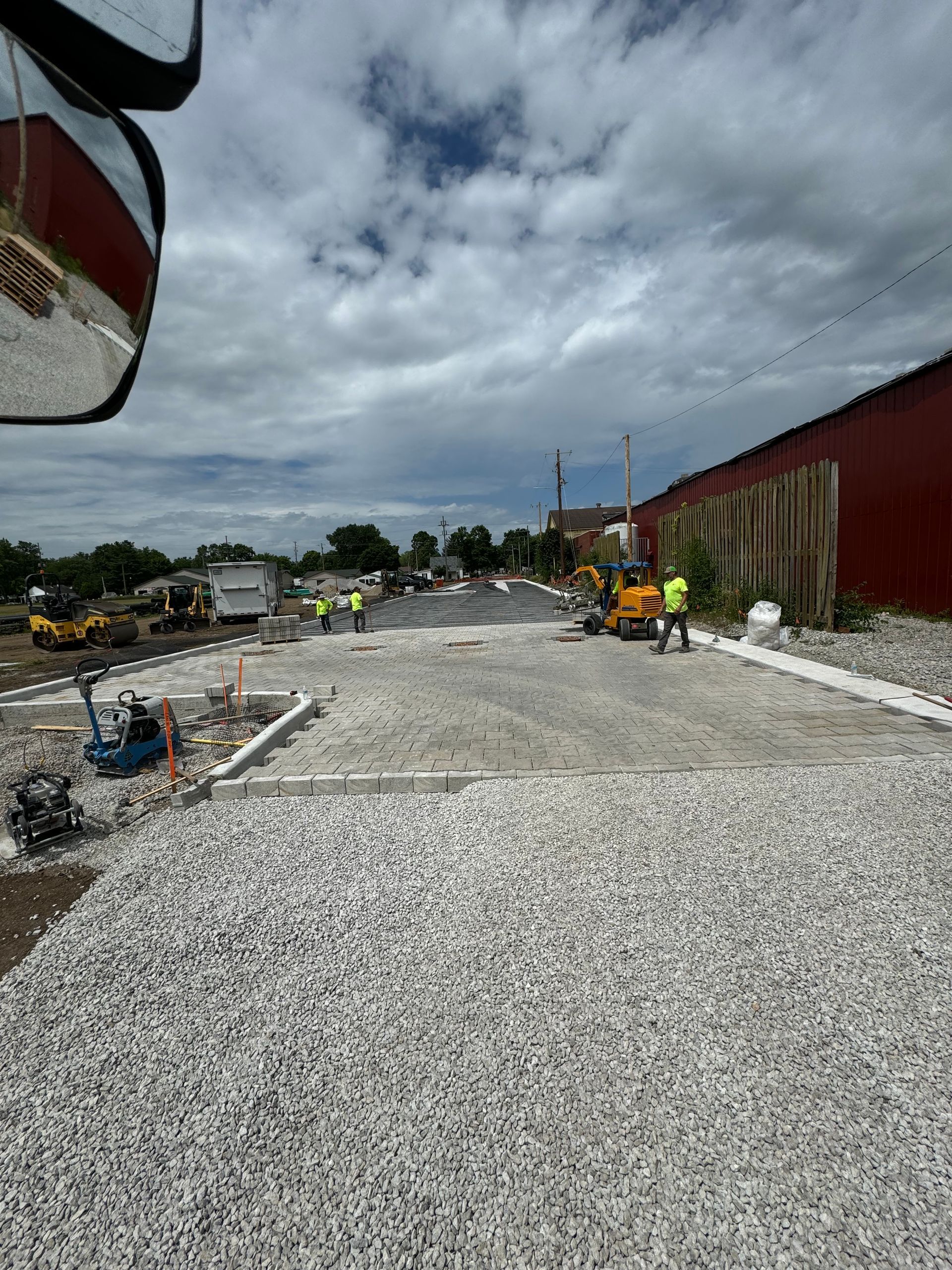 Construction site with gravel road, workers, machinery, and a red building under cloudy skies