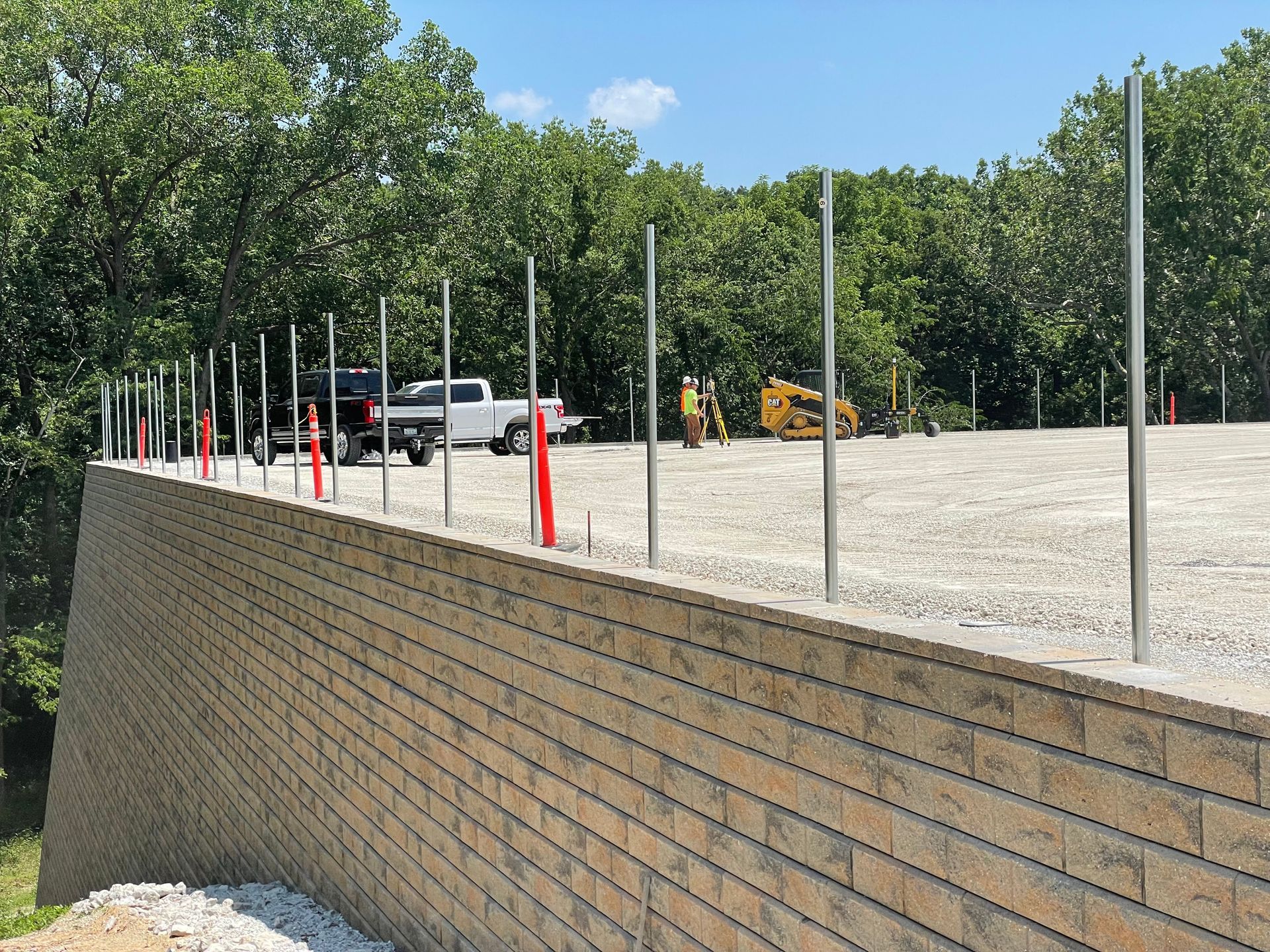 Construction site with retaining wall, gravel lot, parked truck, and tall metal posts against trees.