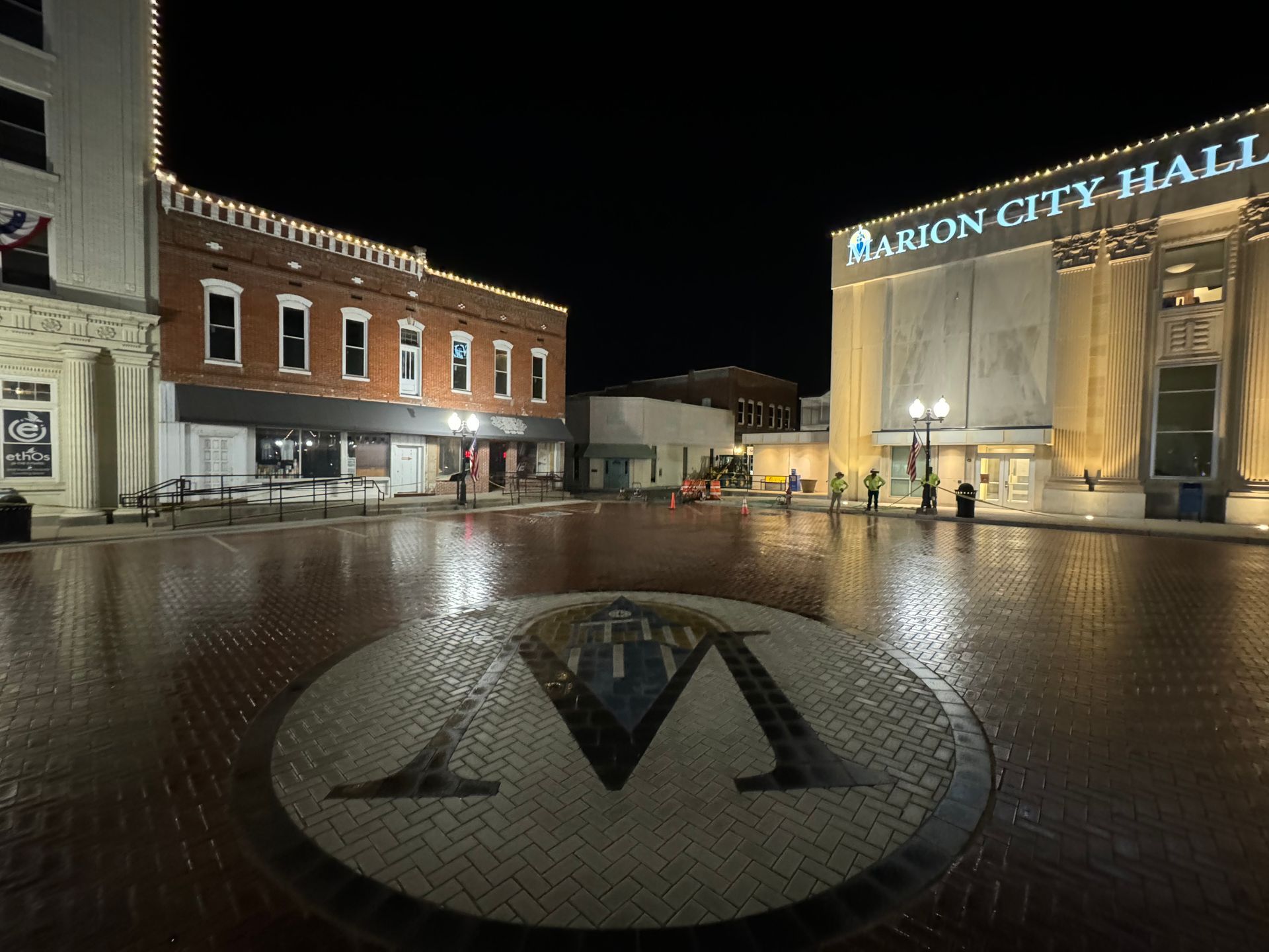 Night view of a wet plaza beside Albion City Hall, with lit buildings and a circular mosaic in the foreground