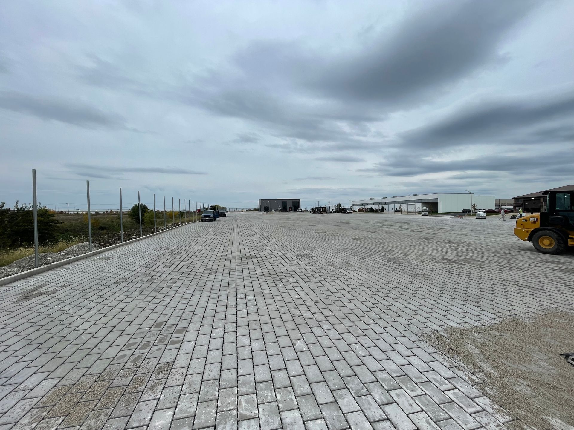 Wide empty cobblestone lot under cloudy sky, with parked construction vehicles in the distance.