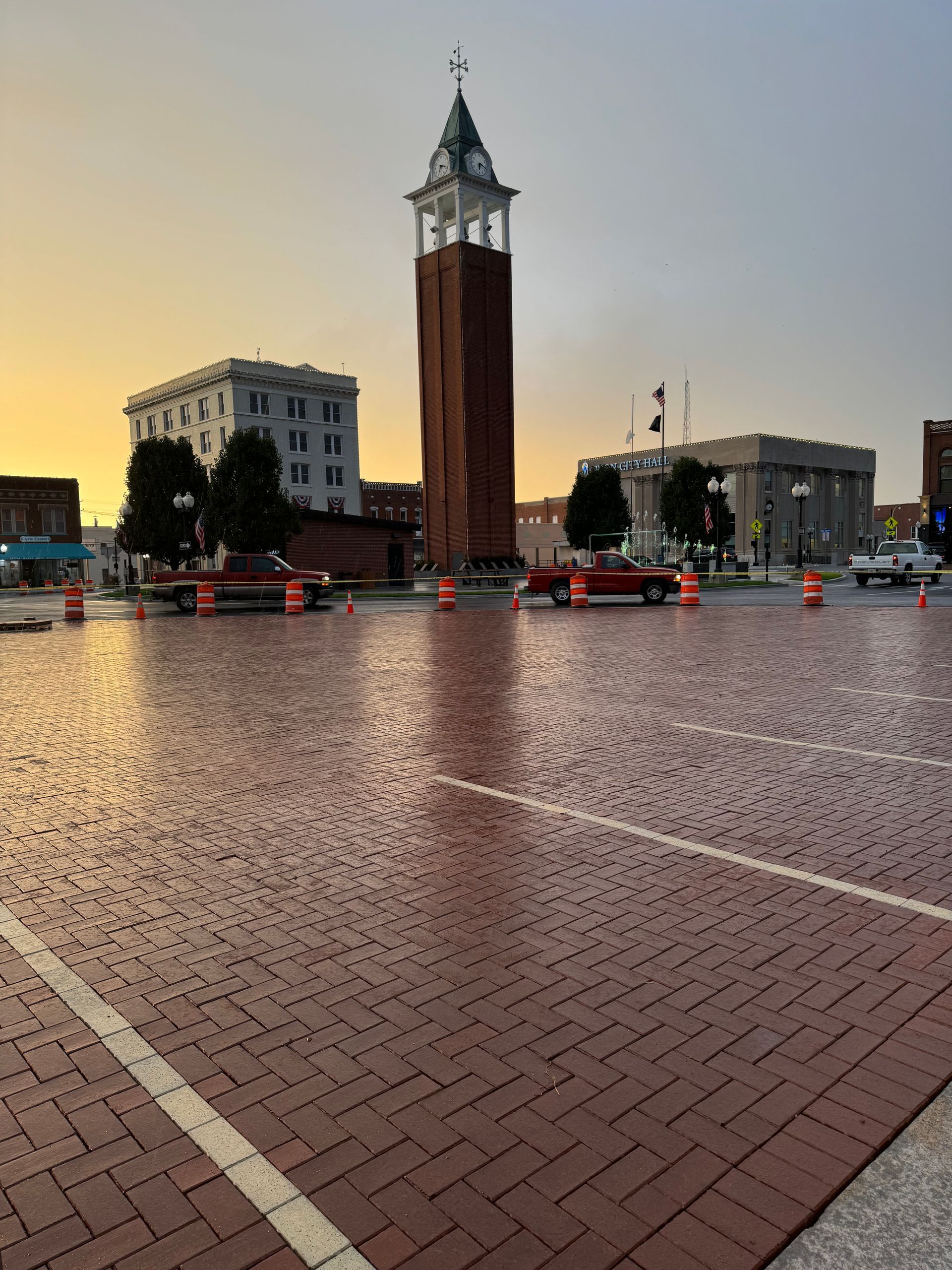 Wet brick plaza with a tall clock tower at sunset, flanked by buildings and a few cars.