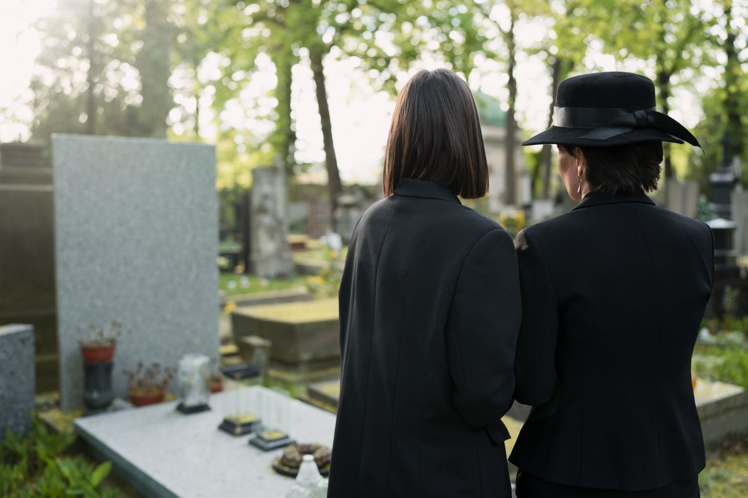 Two people in black clothing stand by a cemetery grave, facing a memorial in sunlight.
