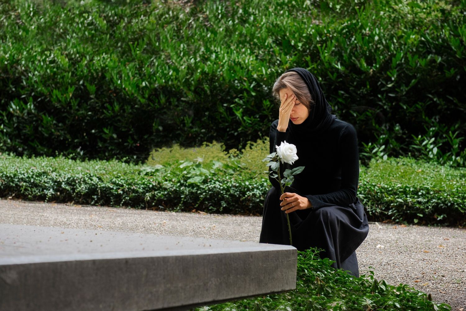 Person kneels in a garden beside a stone bench, holding a white flower and looking down.