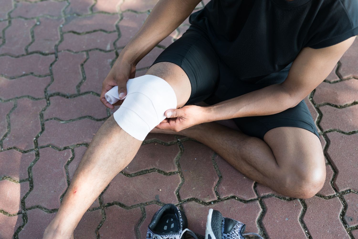 Person wrapping a white bandage around a knee while sitting on brick pavement, with shoes nearby