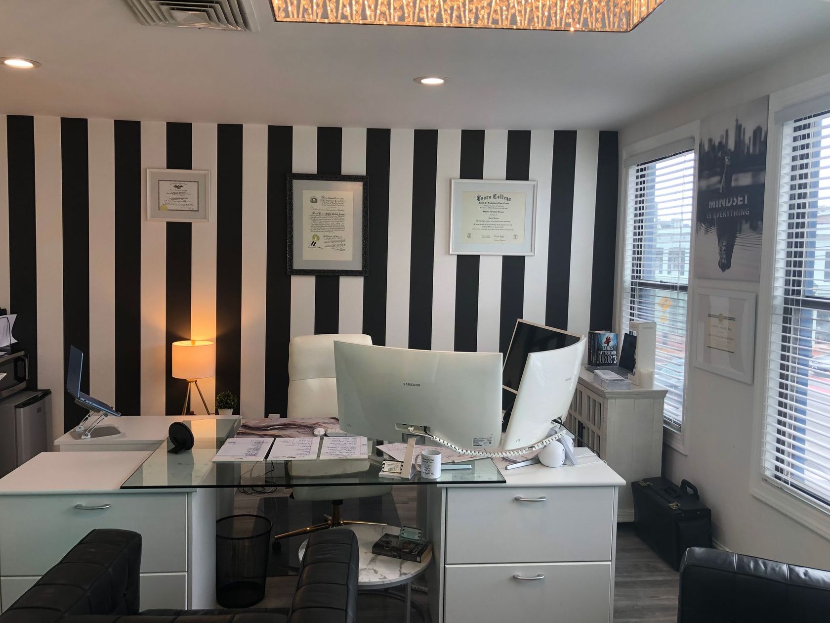 Office desk with computer, striped black-and-white wall, framed certificates, and window blinds