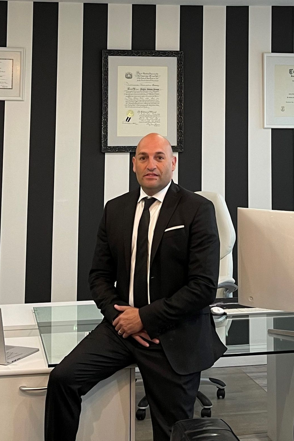Man in a black suit sitting on a glass desk in an office with black-and-white striped walls.