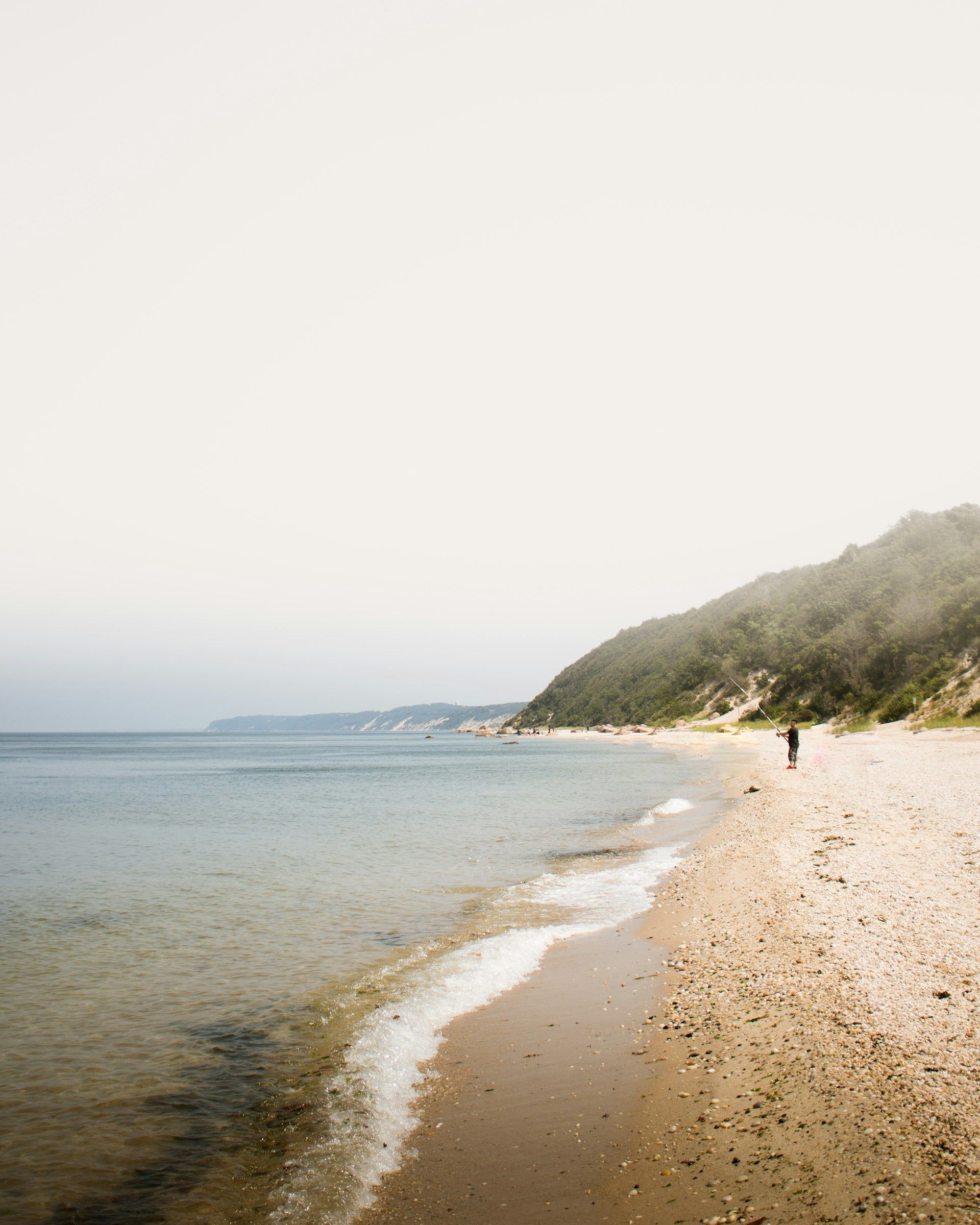 Sandy beach with calm water, a distant shoreline, and a lone person walking near the coast