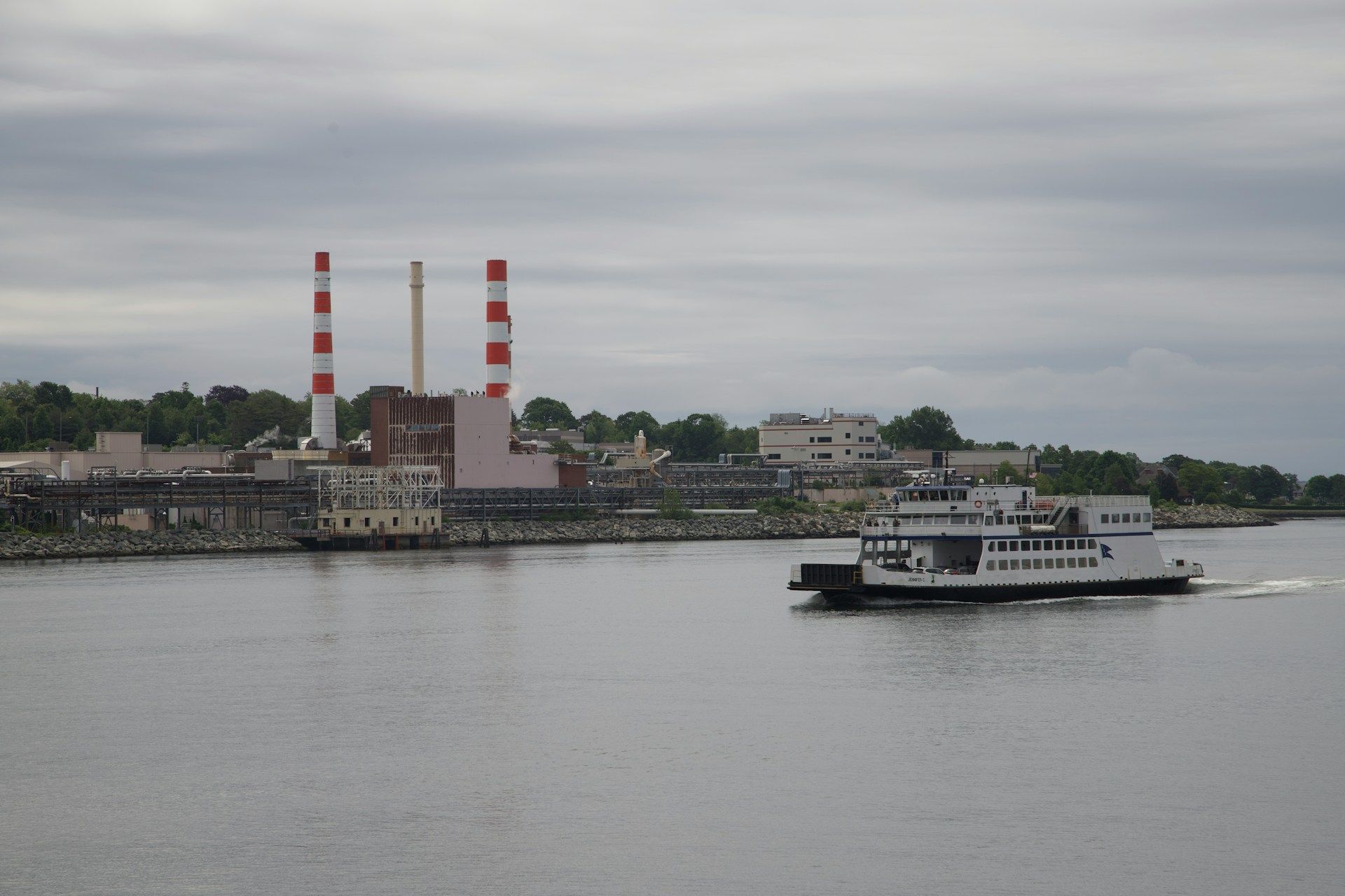 Ferry on a gray river with a striped smokestack and industrial shoreline in the background