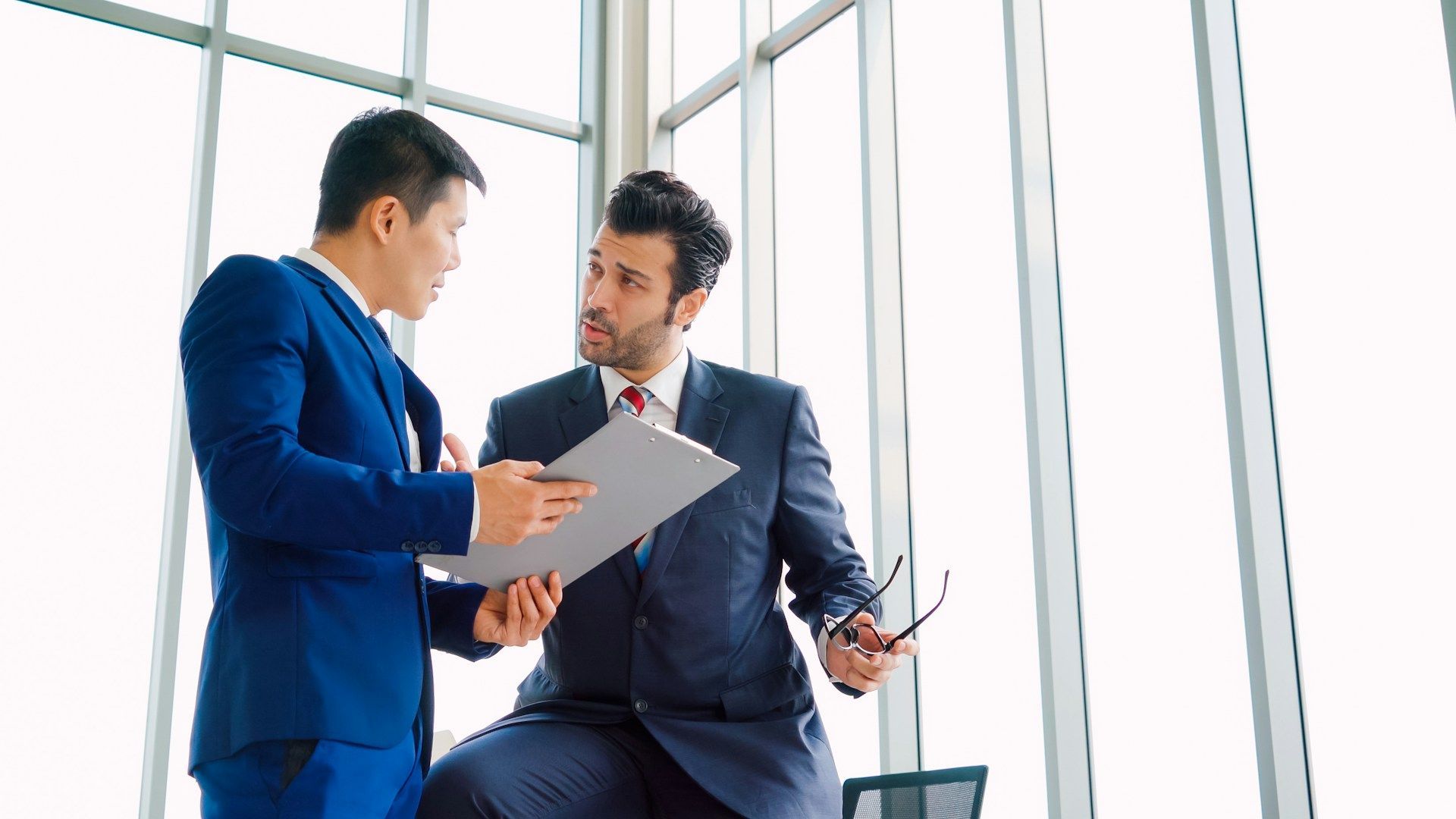 Two businesspeople in suits discussing a document in a bright office with large windows.