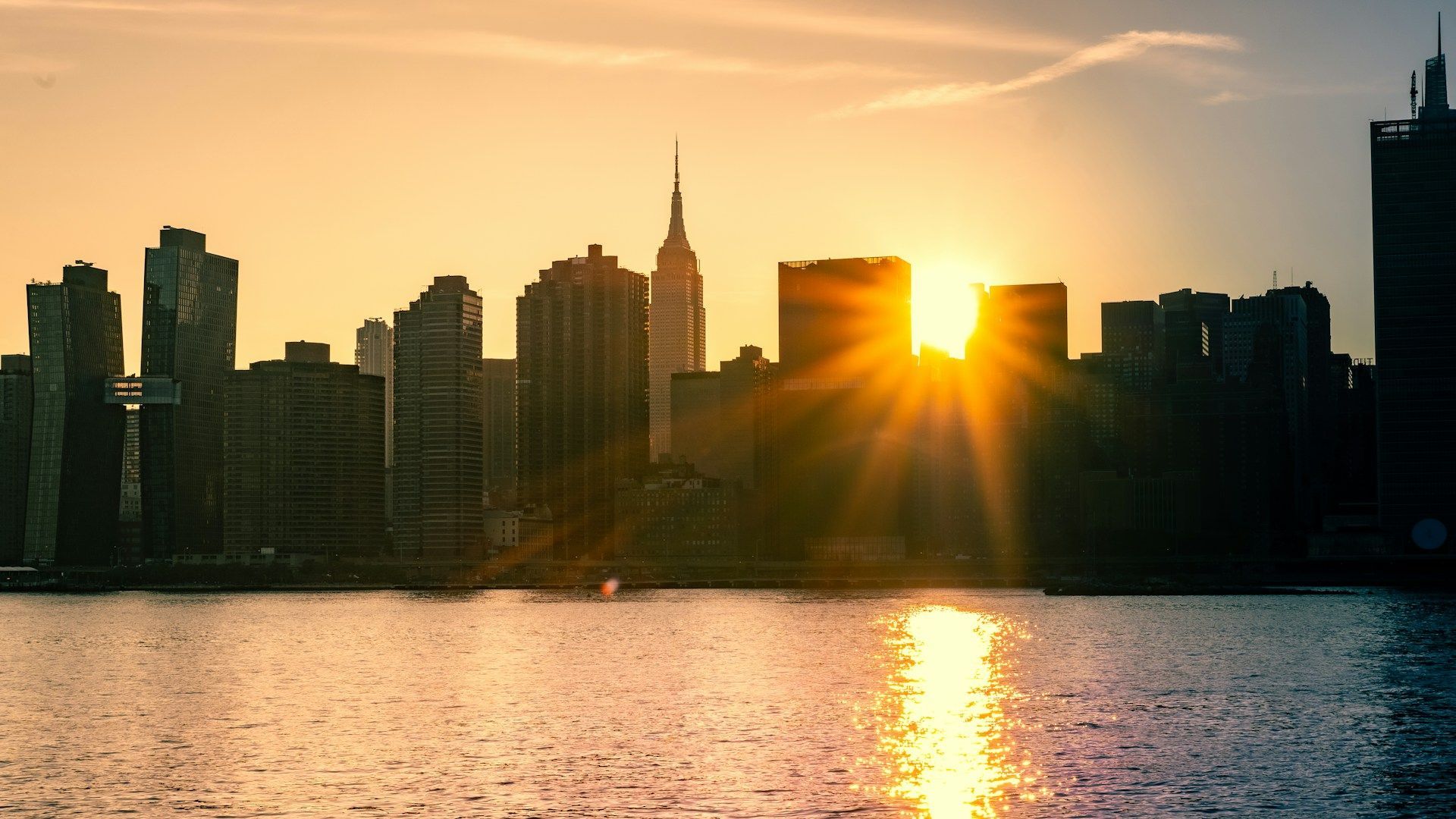 Sunset over a city skyline, with tall buildings silhouetted above shimmering water