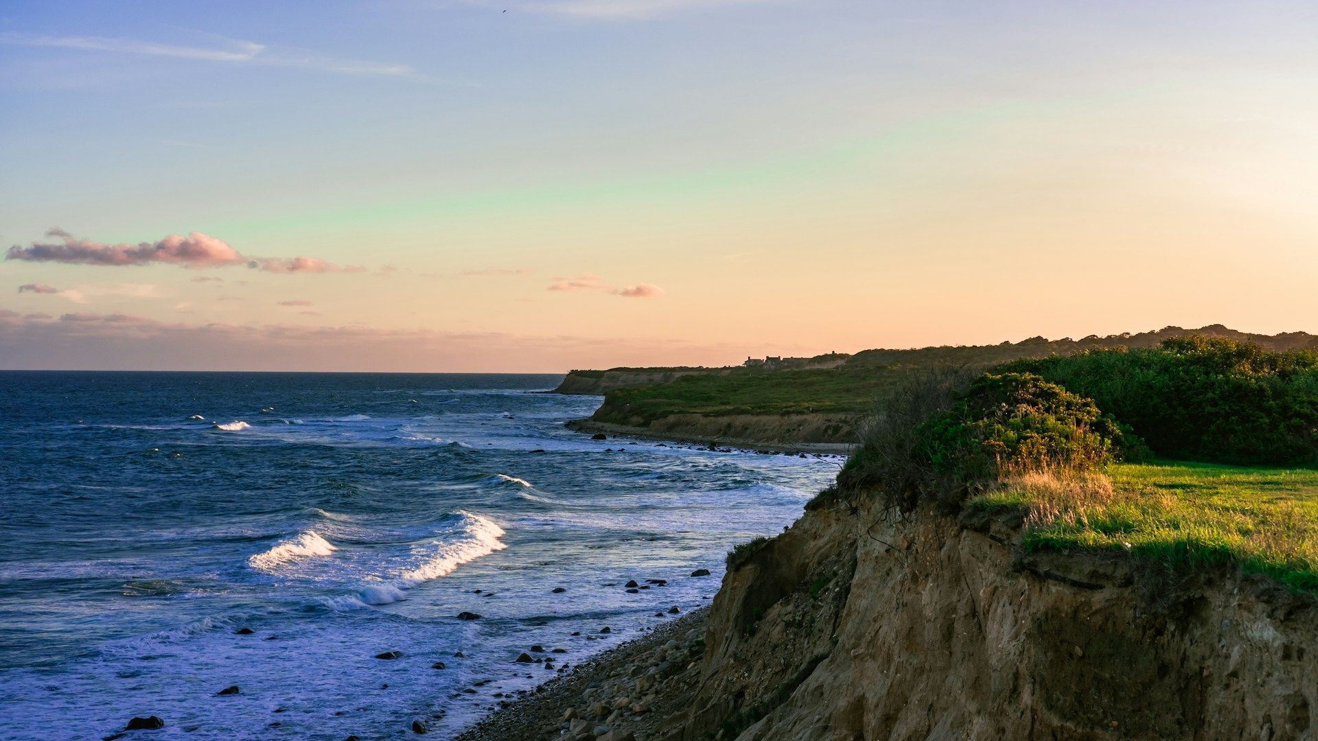 Rocky seaside cliffs beside choppy blue water under a pastel sunset sky