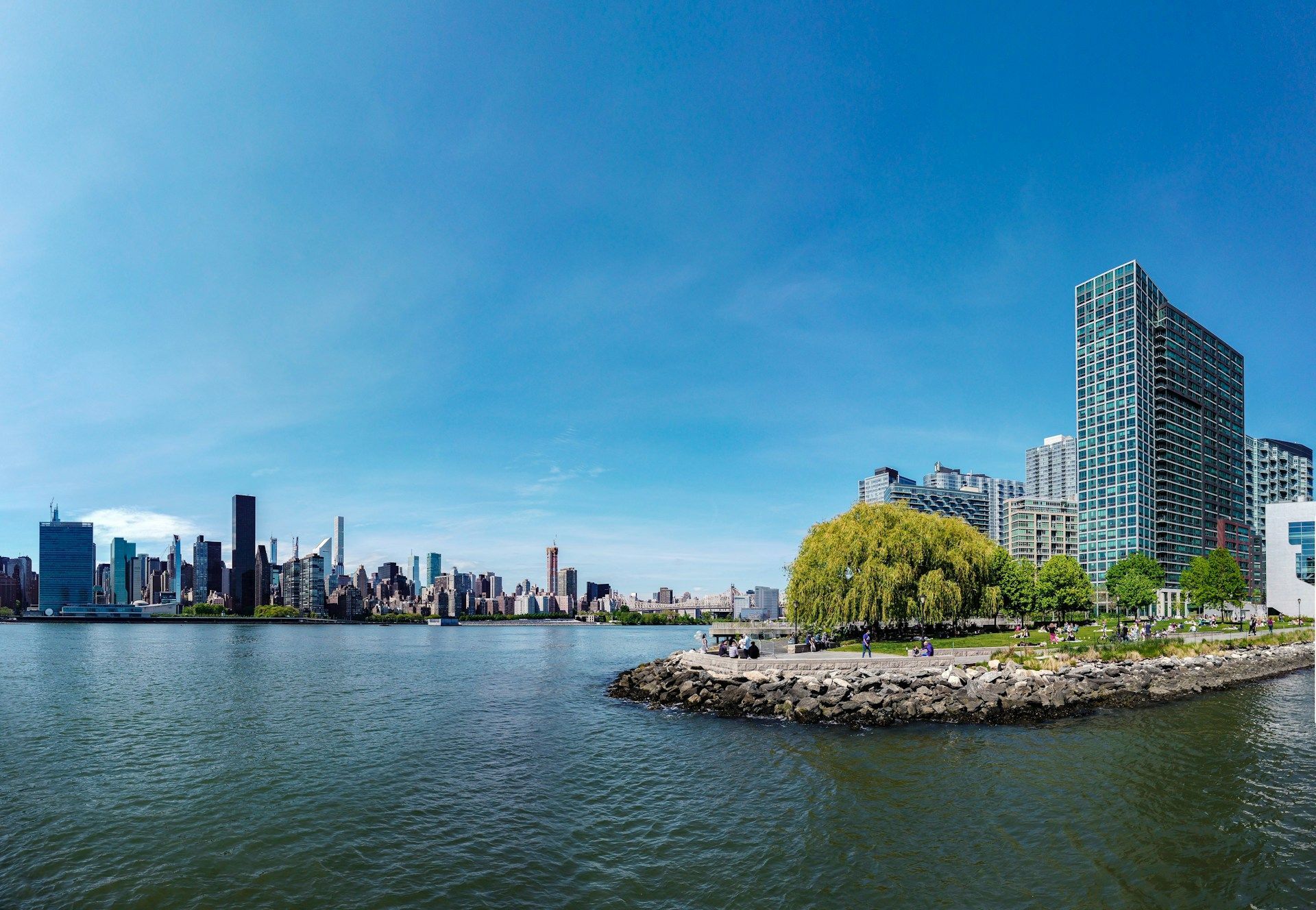 City skyline across a river with a rocky green shoreline and tall blue glass buildings under a clear sky