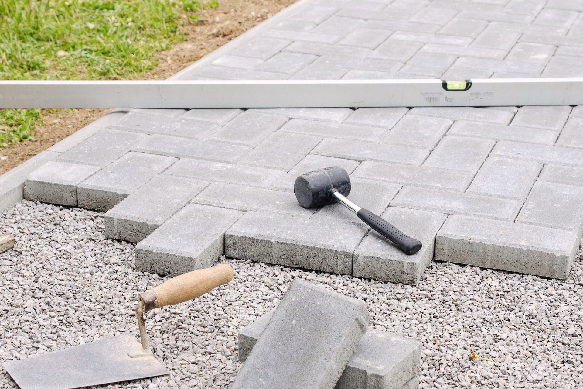 Gray paving stones being laid on gravel with a spirit level, mallet, and trowel nearby.