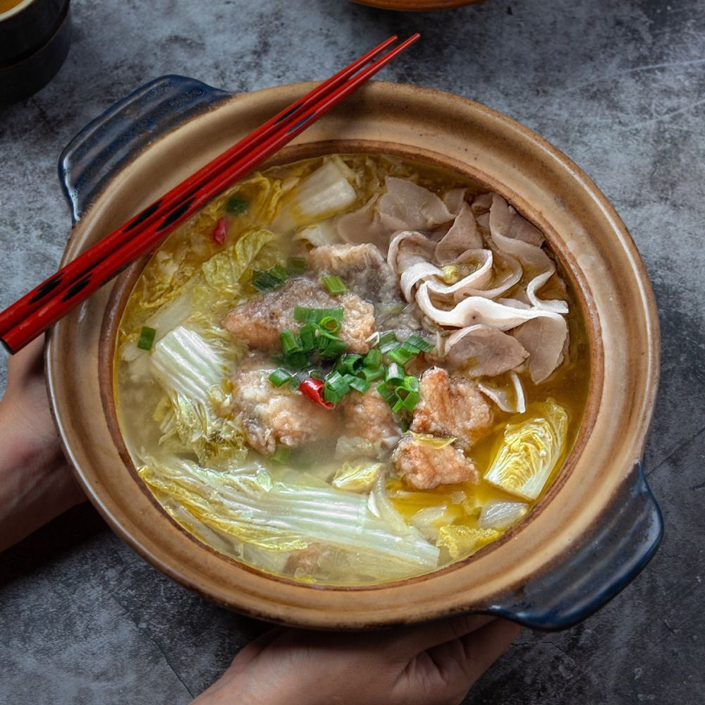 雙手捧著一口盛有魚、豬肉和蔬菜的中國湯的陶罐，上面放著筷子。