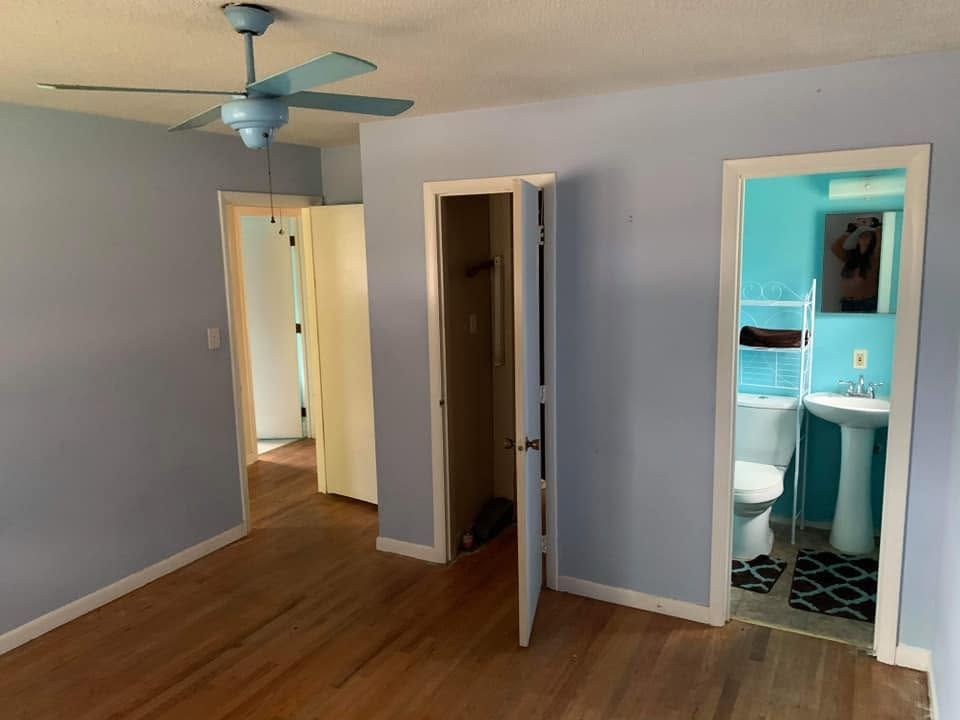 Empty bedroom with light blue walls, hardwood floor, ceiling fan, and open doors to a hallway and bathroom.