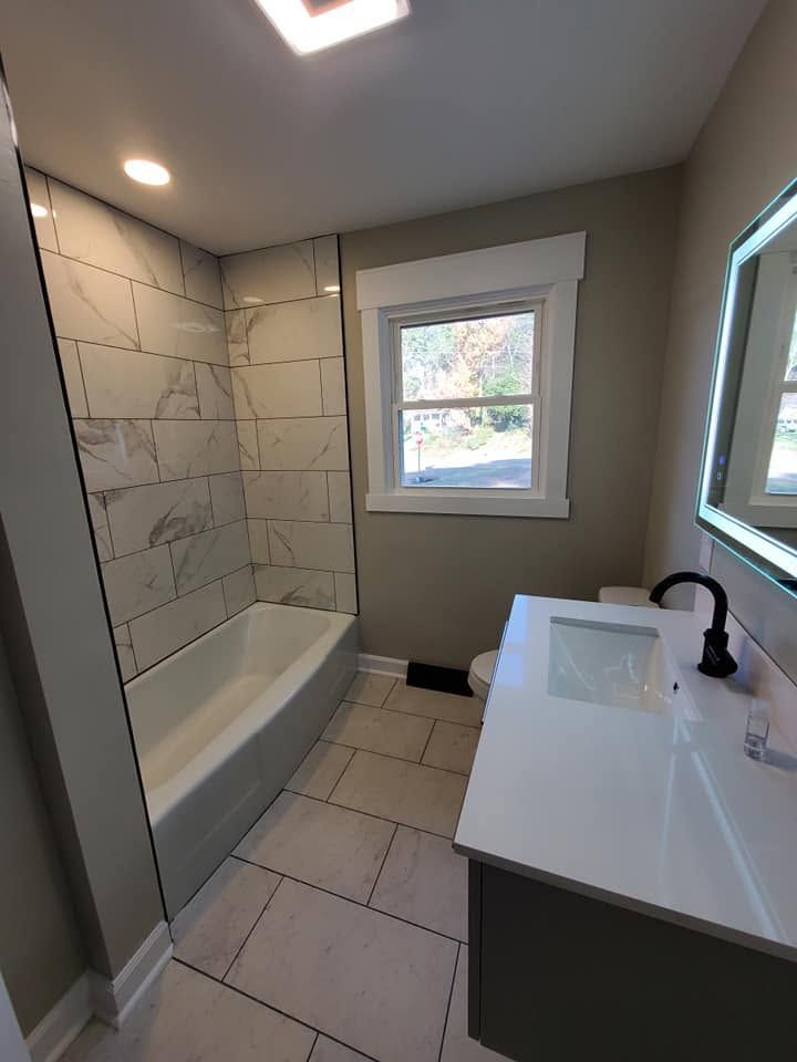 Small modern bathroom with tiled shower-tub, white vanity, and window.