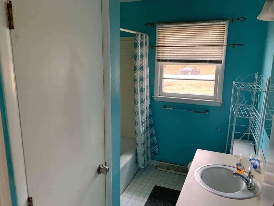 Small teal bathroom with a shower, window blinds, white sink, and a wall-mounted drying rack.