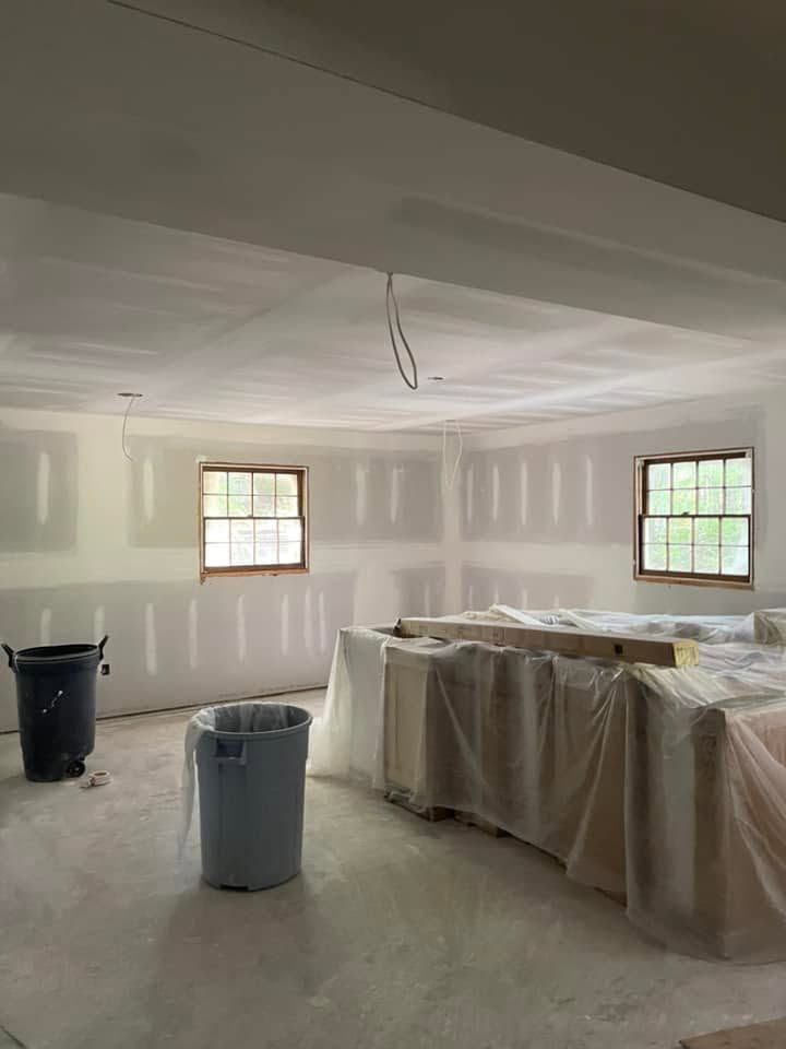 Unfinished room under renovation with drywall, covered furniture, two windows, and paint buckets on the floor.