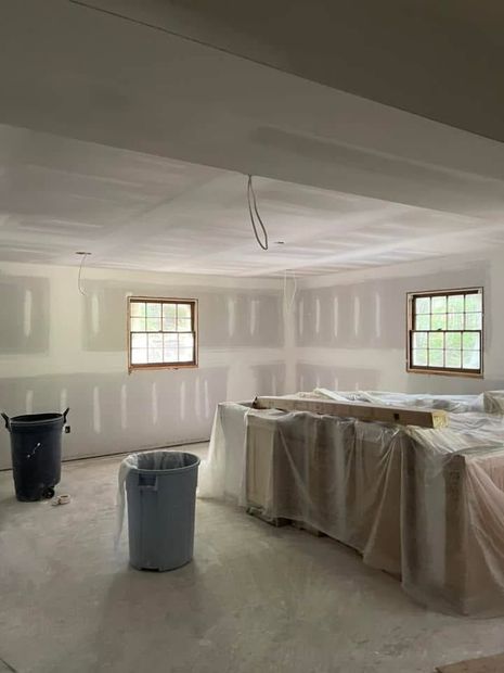 Unfinished room under renovation with drywall, covered furniture, two windows, and paint buckets on the floor.