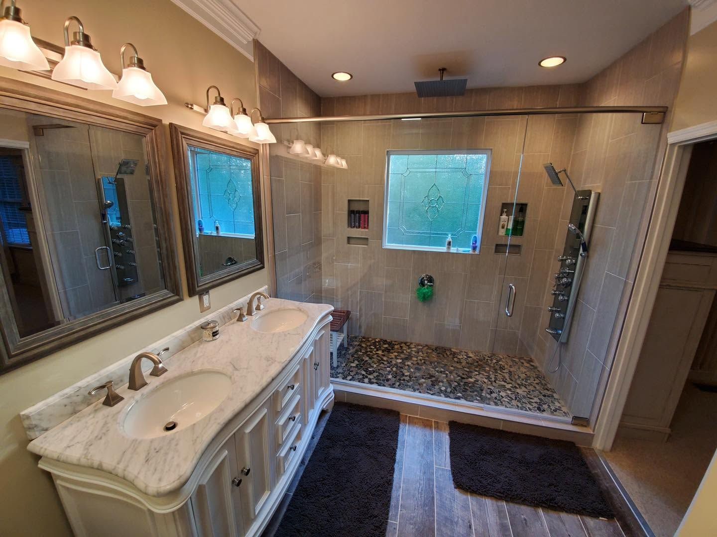 Spacious bathroom with double vanity, large mirrors, glass shower, and dark rug on tiled floor.