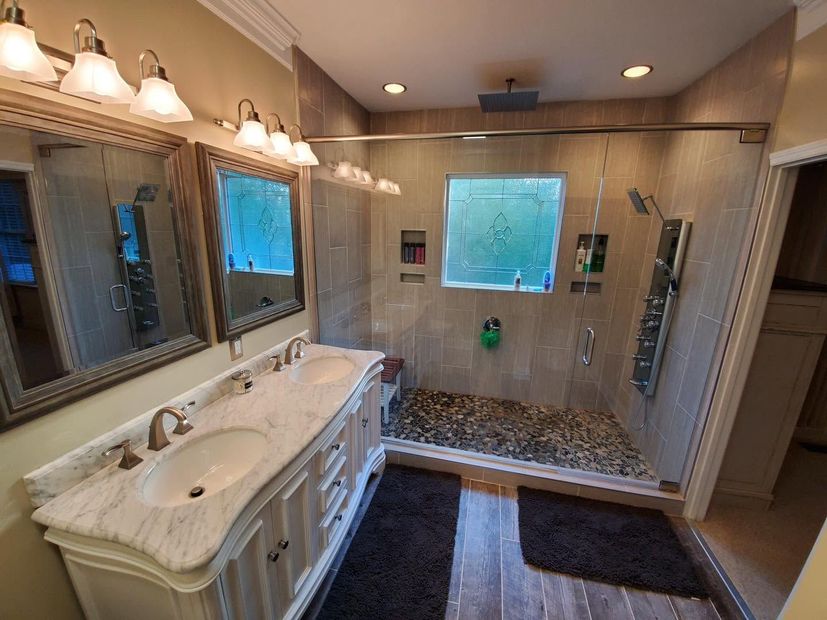 Spacious bathroom with double vanity, large mirrors, glass shower, and dark rug on tiled floor.