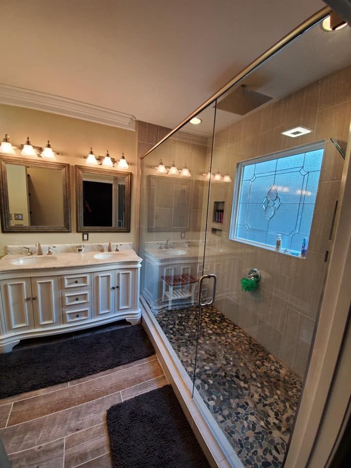 Bathroom with double vanity, mirror lights, and glass shower with pebble floor and tiled walls.