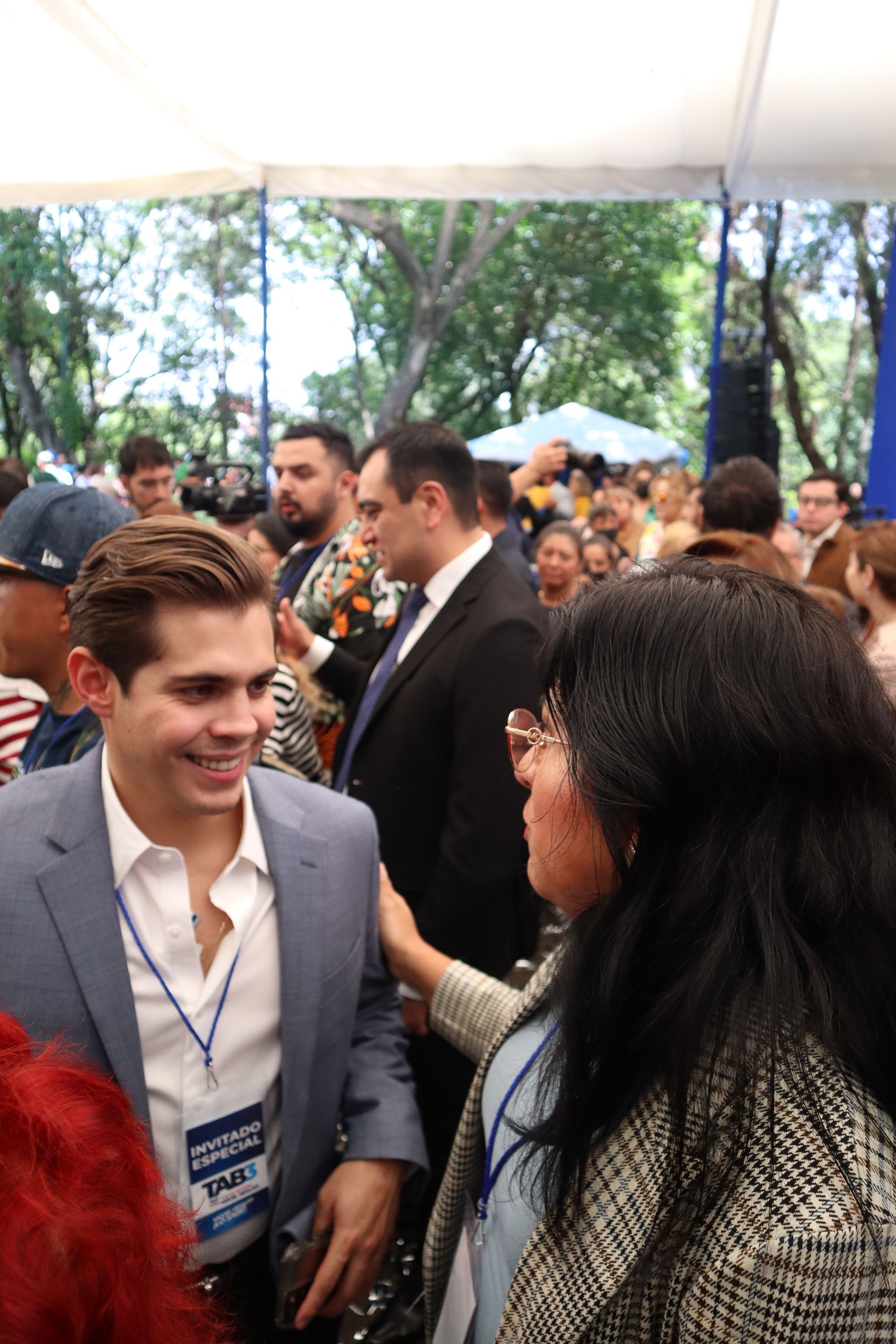Two people talking at an outdoor event, with a crowd and trees in the background.