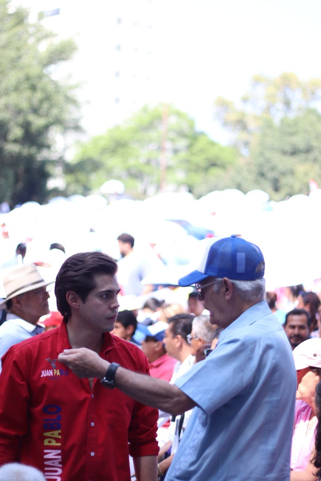 Man in red shirt speaking to elderly man in blue cap at an outdoor crowd event