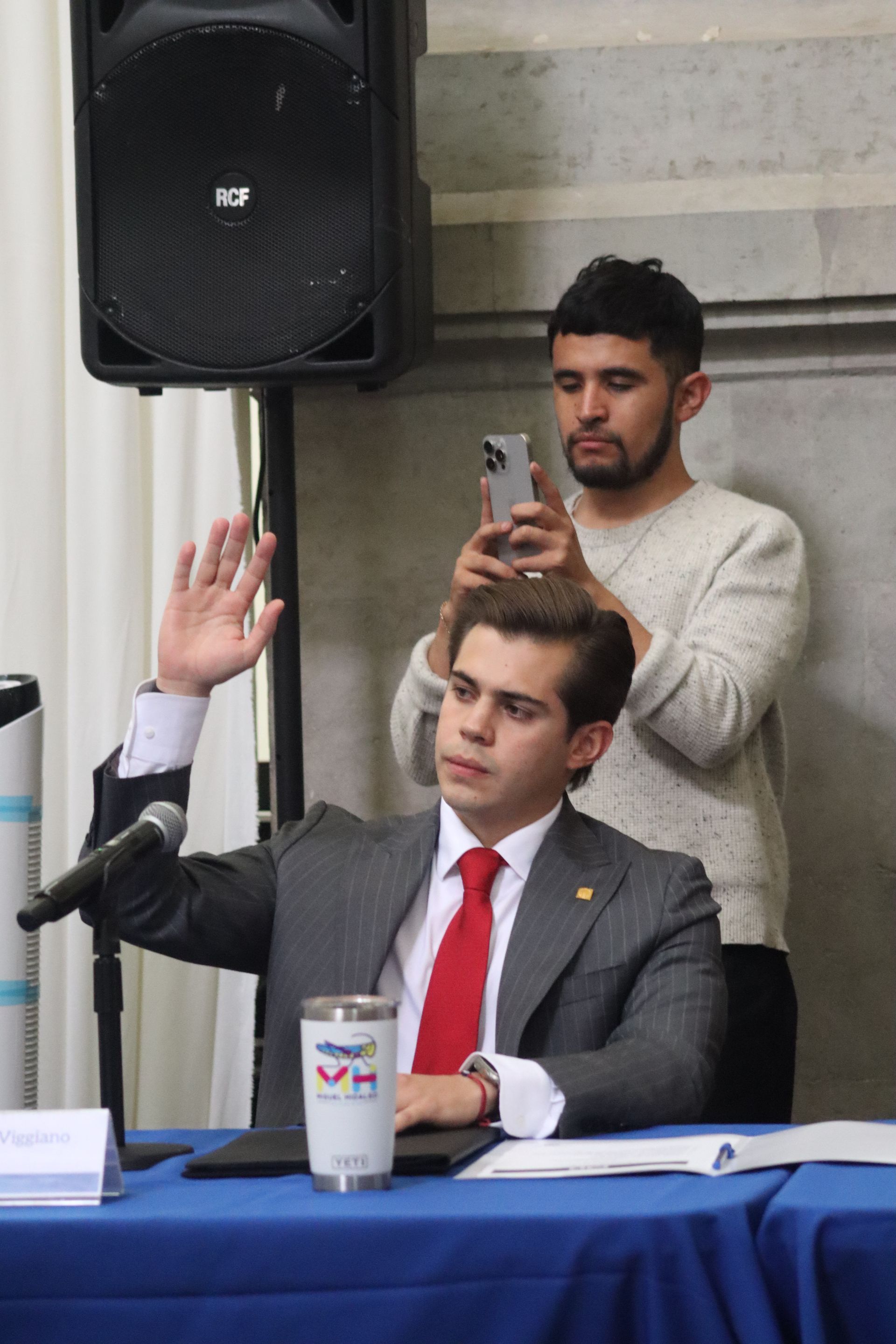 Seated man in a gray suit raises his hand at a table while another man stands behind filming with a phone.