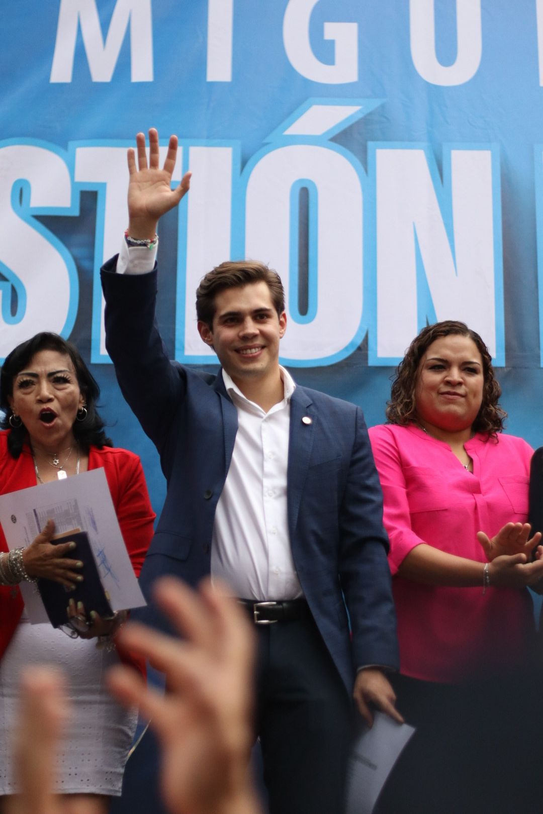 Man in a navy suit waves onstage beside two women before a blue campaign backdrop