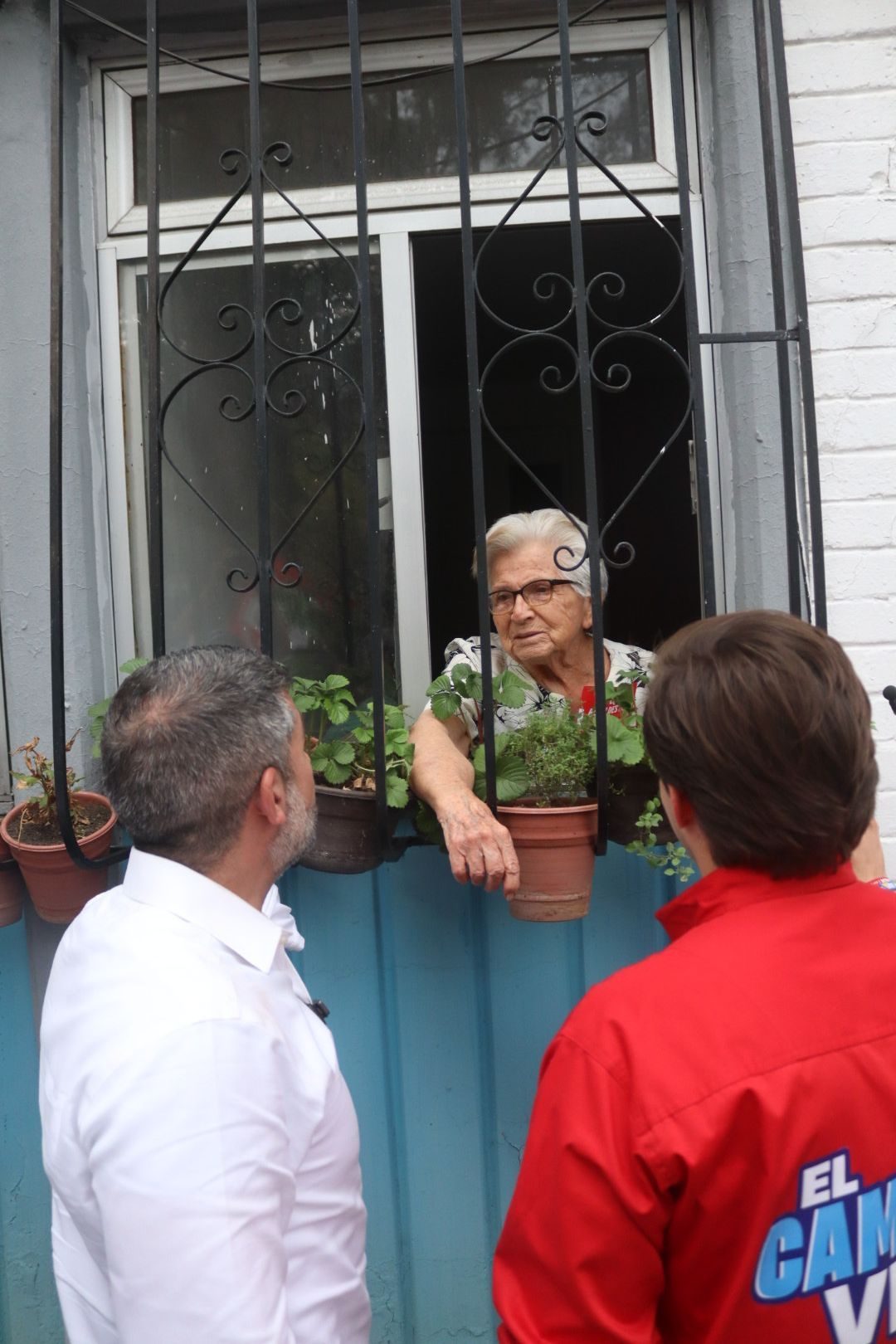 Older woman leaning at a window, talking to two people outside a blue house with metal bars.