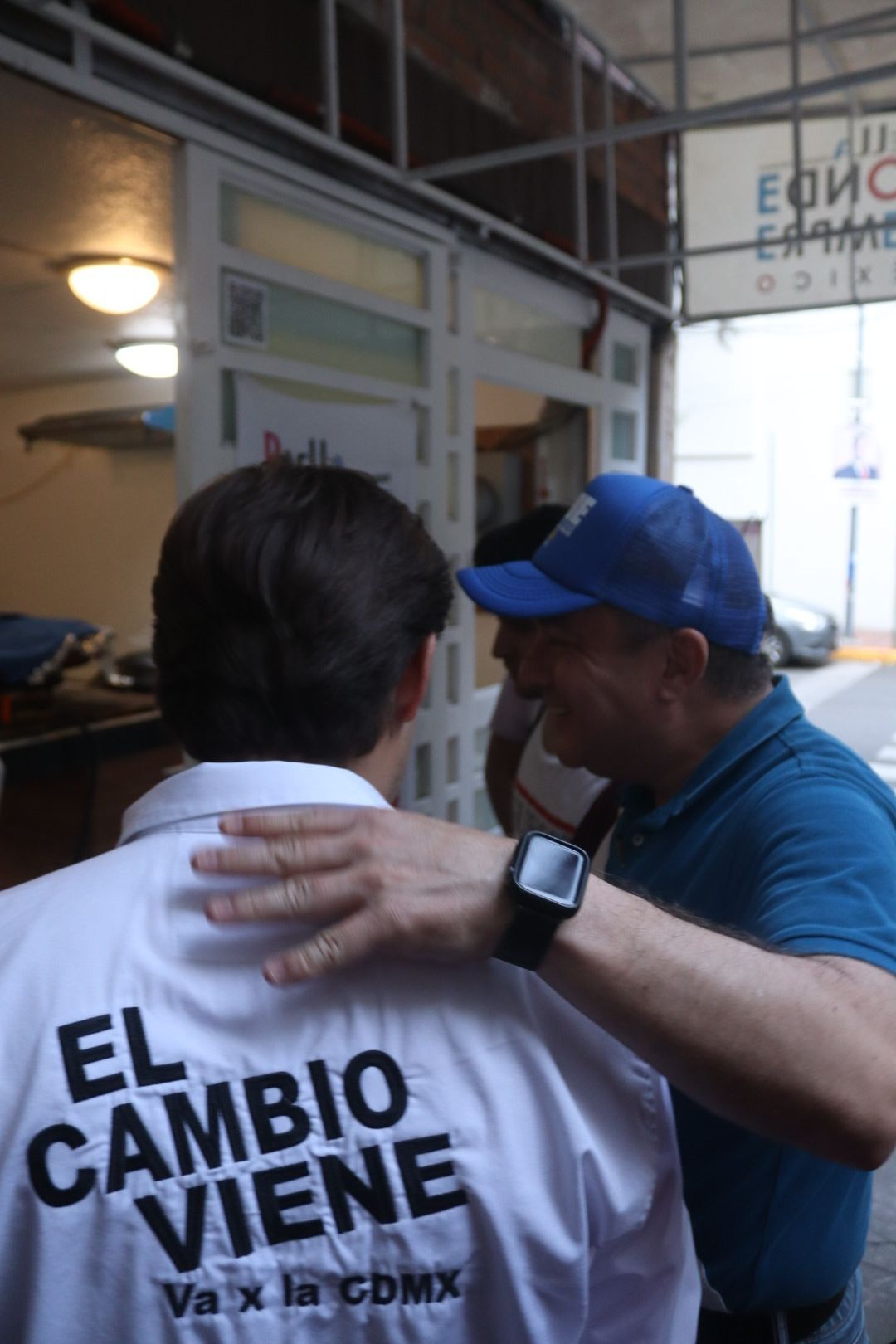 Two men talking indoors, one in a blue cap patting the other’s back near a window.