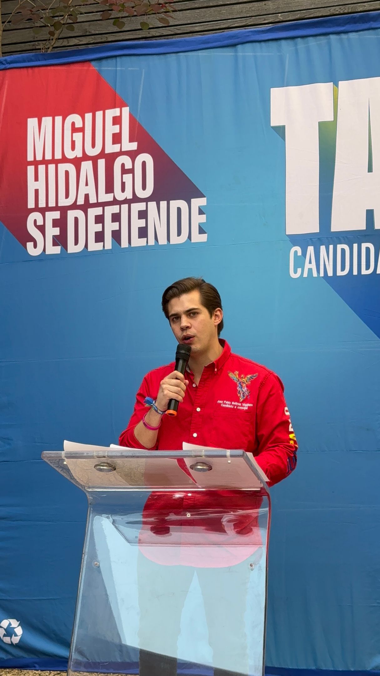 Man speaking at a podium during a political event, with red and blue campaign banners behind him.