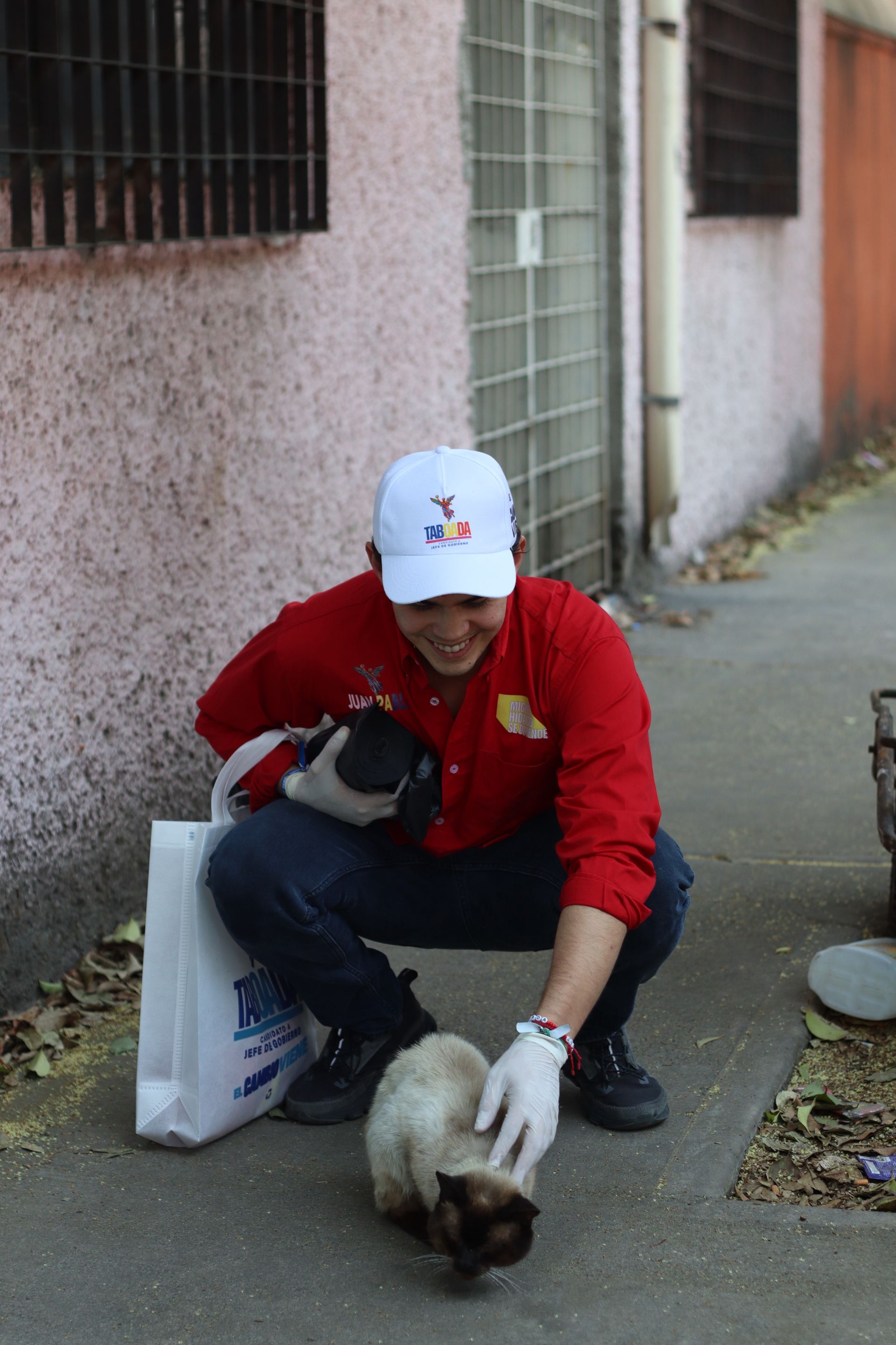 Person in red jacket crouches on sidewalk petting a cat beside a building