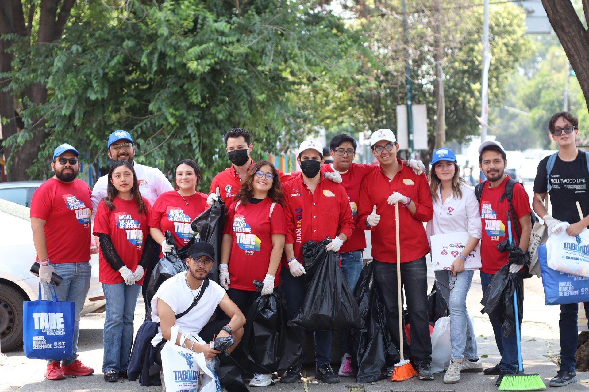 Group of volunteers in red shirts posing outdoors with brooms and trash bags during a community cleanup