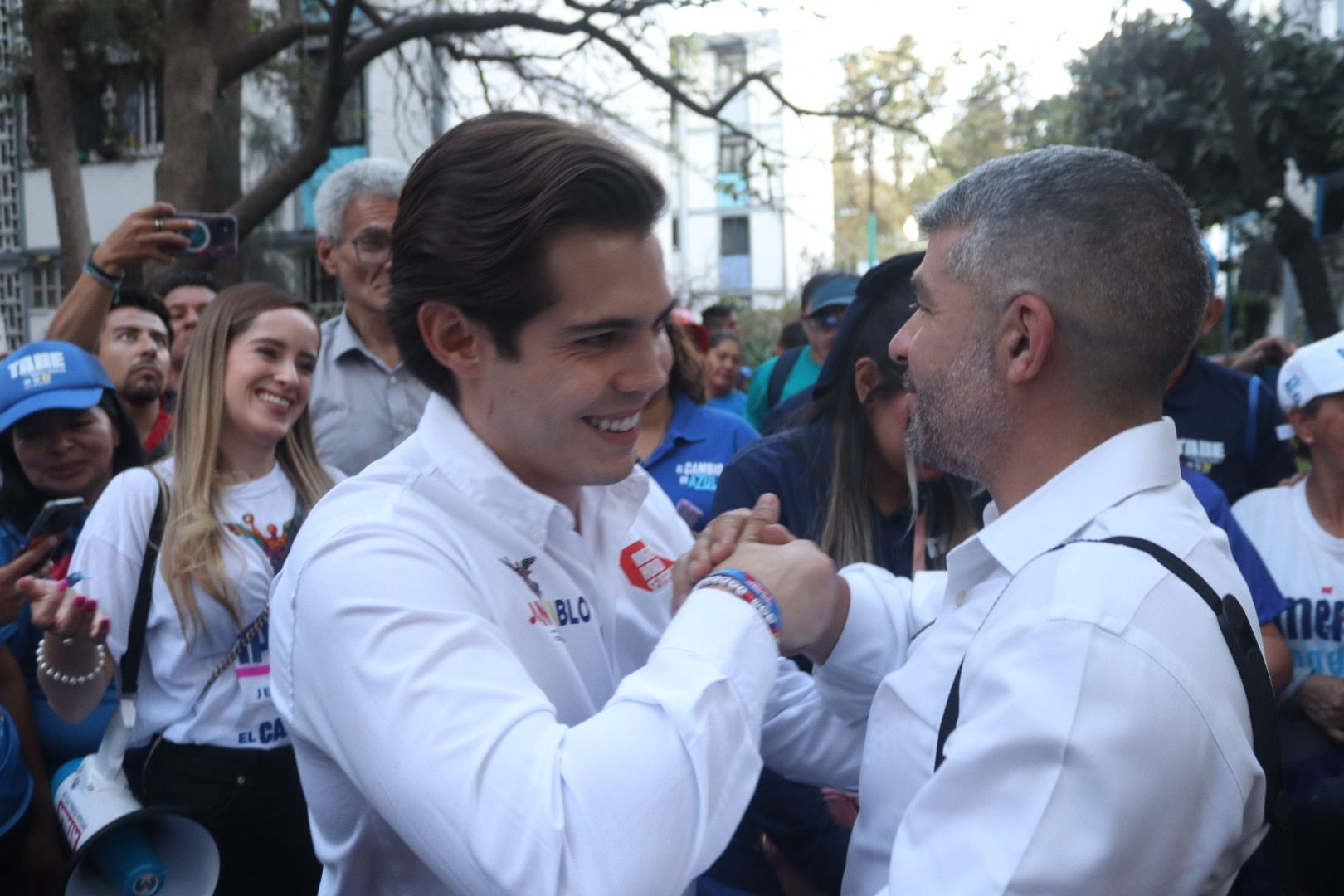 Two men in white shirts smile and clasp hands at a crowded outdoor event
