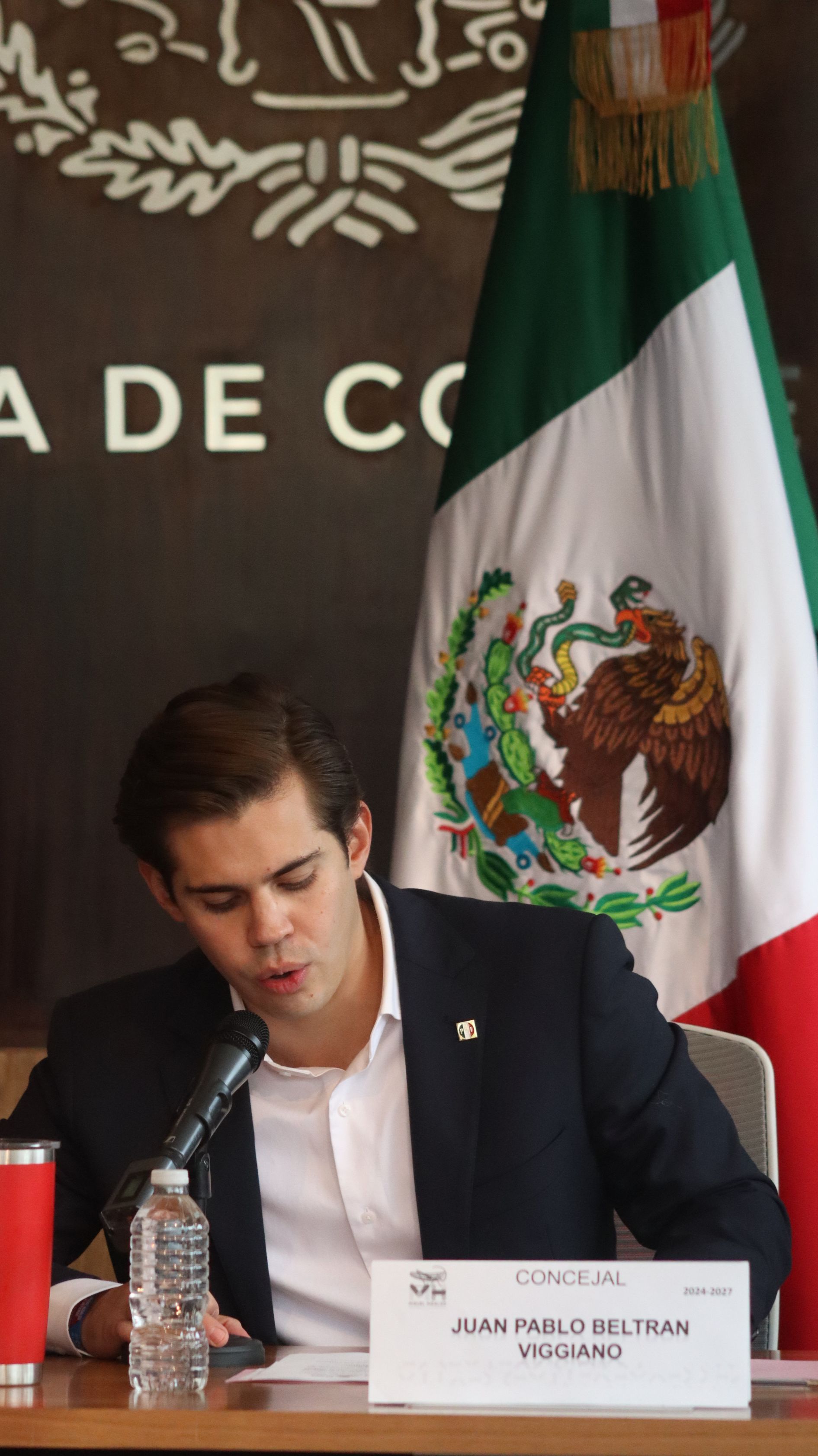 Man speaking at a podium with a Mexican flag in the background and a nameplate in front