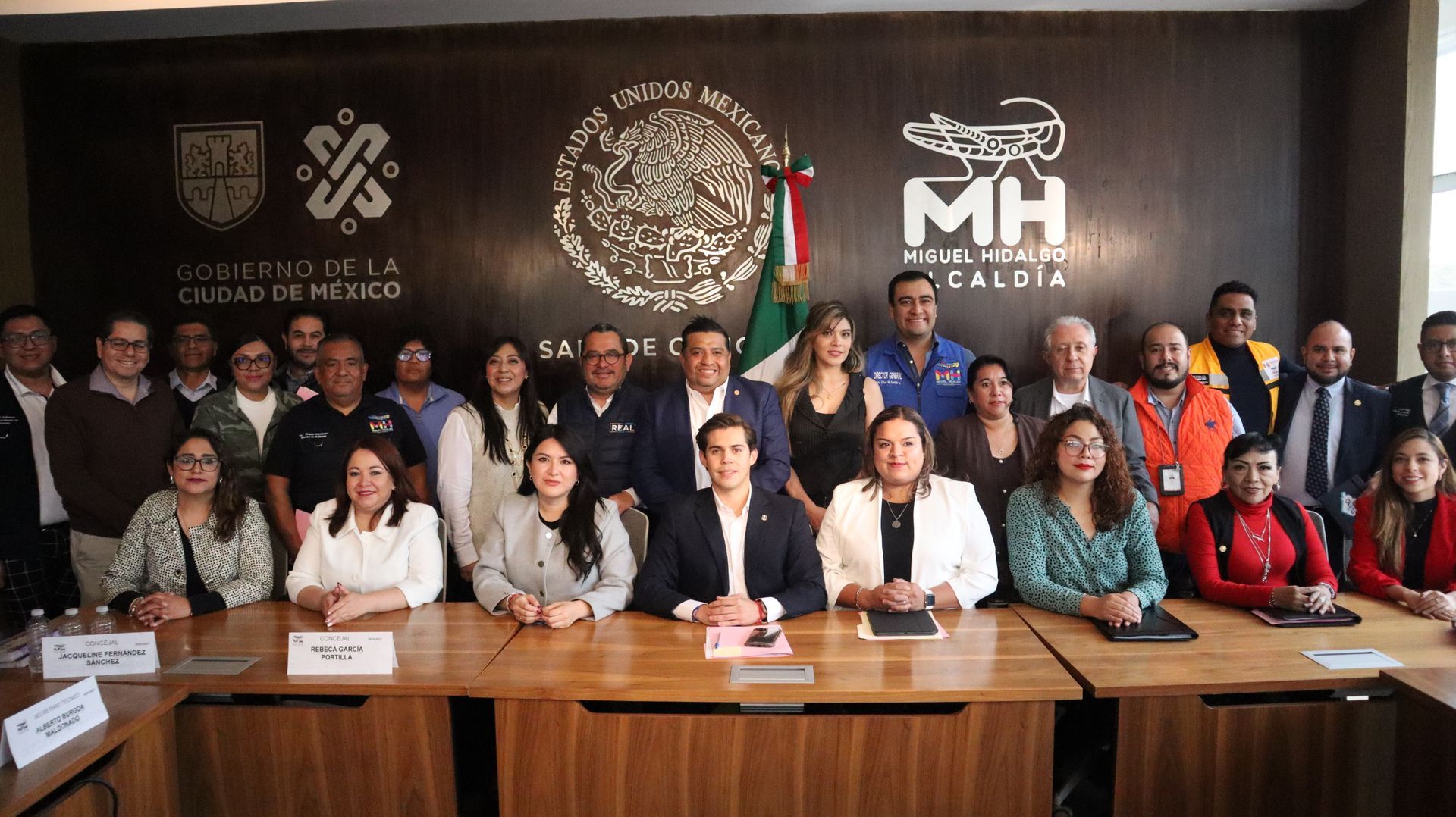 Group portrait of people seated and standing in a conference room, with a dark wall logo backdrop behind them
