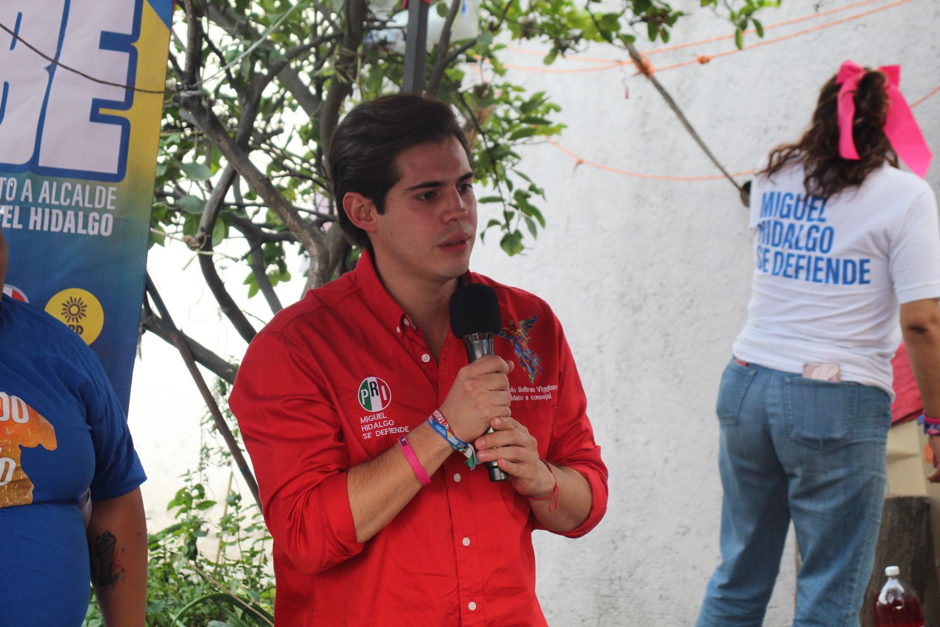Man in a red shirt speaking into a microphone at an outdoor event, with people and a mannequin nearby.