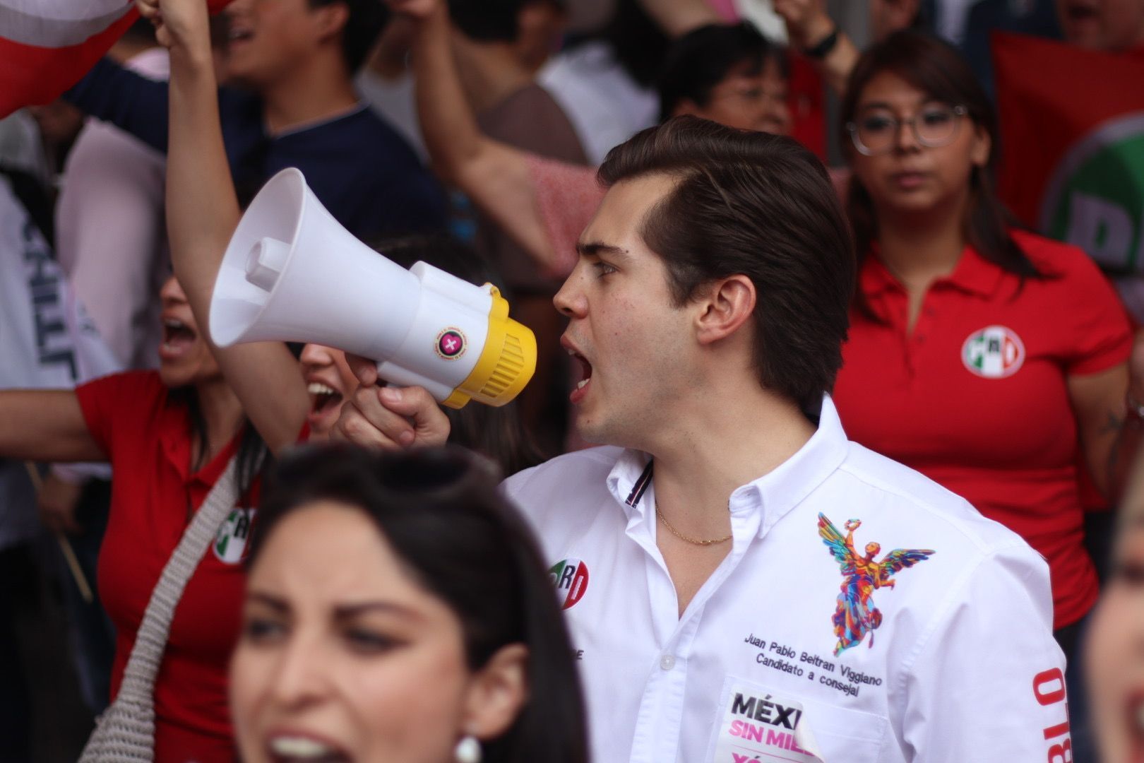 Man in white shirt speaking through a megaphone at a crowd, with people in red shirts behind him.