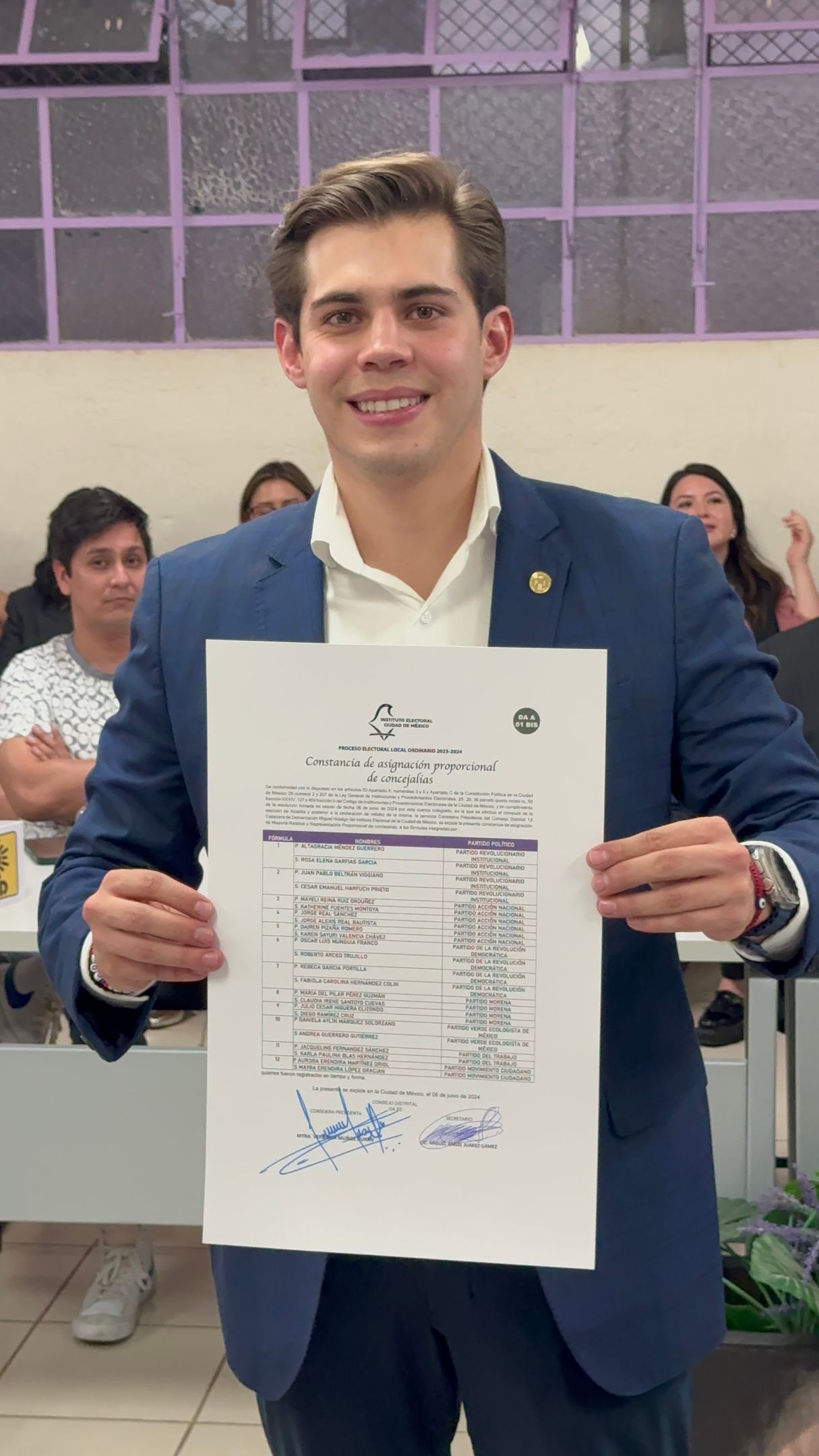 Man in blue suit holding a certificate and smiling indoors, with seated people in the background