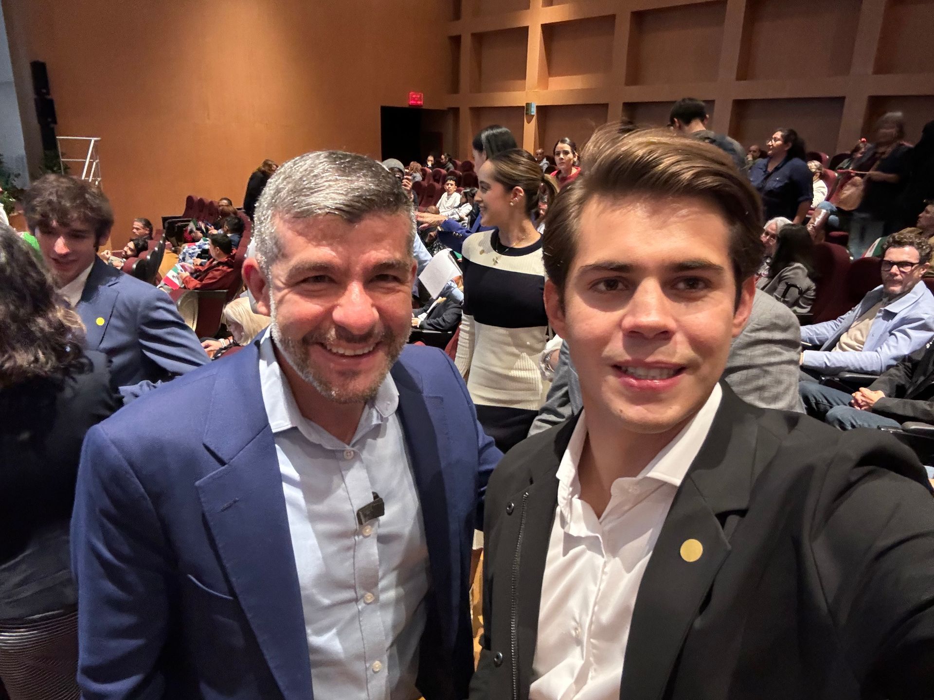 Two men posing for a selfie in a conference hall, with seated attendees in the background.