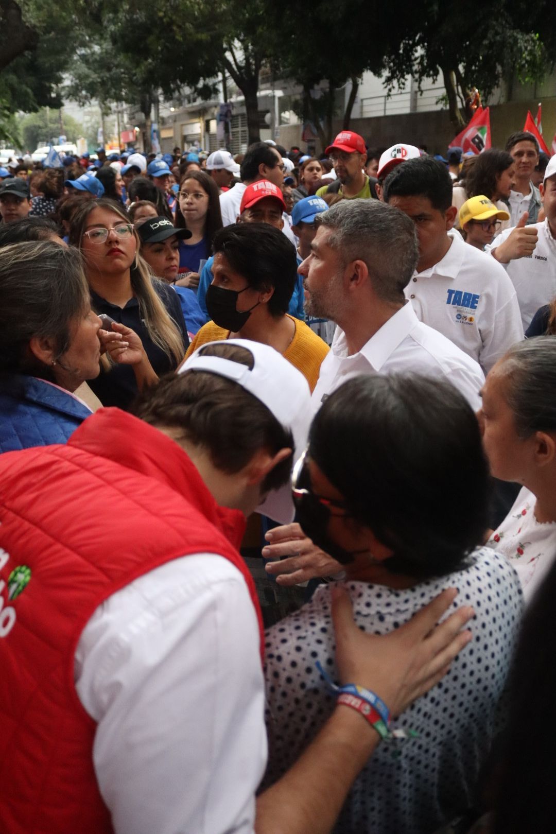 Crowd gathered outdoors at a public event, with people in white and red shirts speaking closely together.