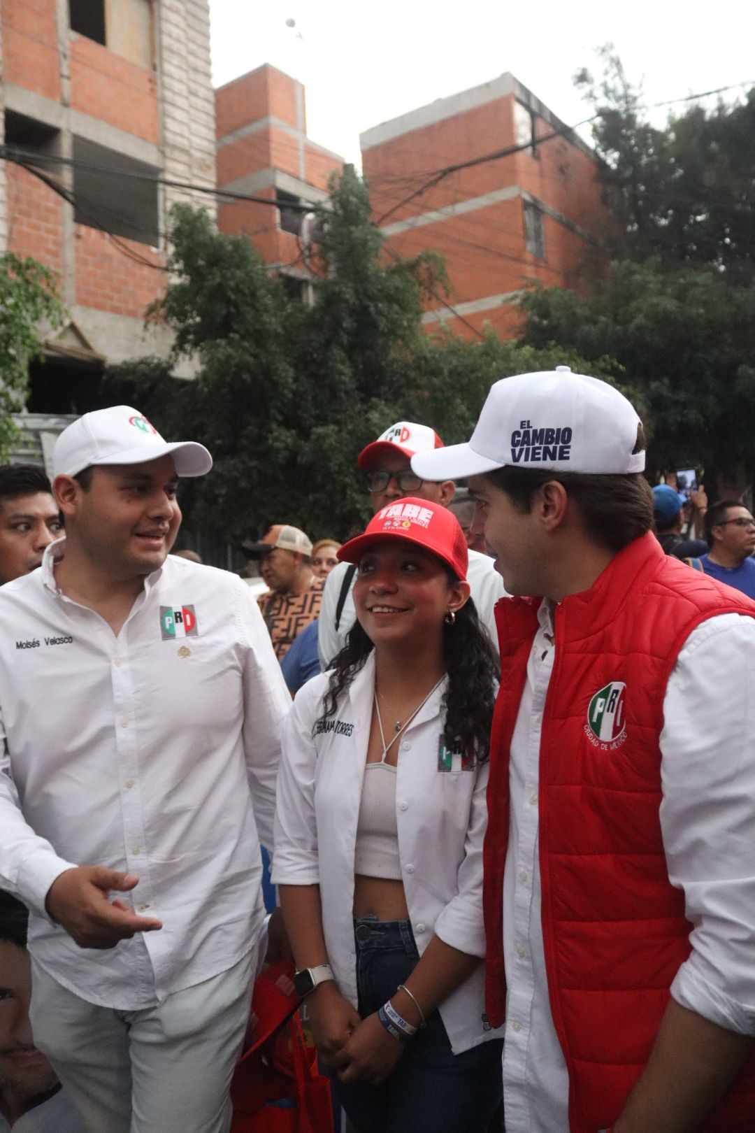 Three people in white and red campaign-style clothing standing outdoors near brick buildings.