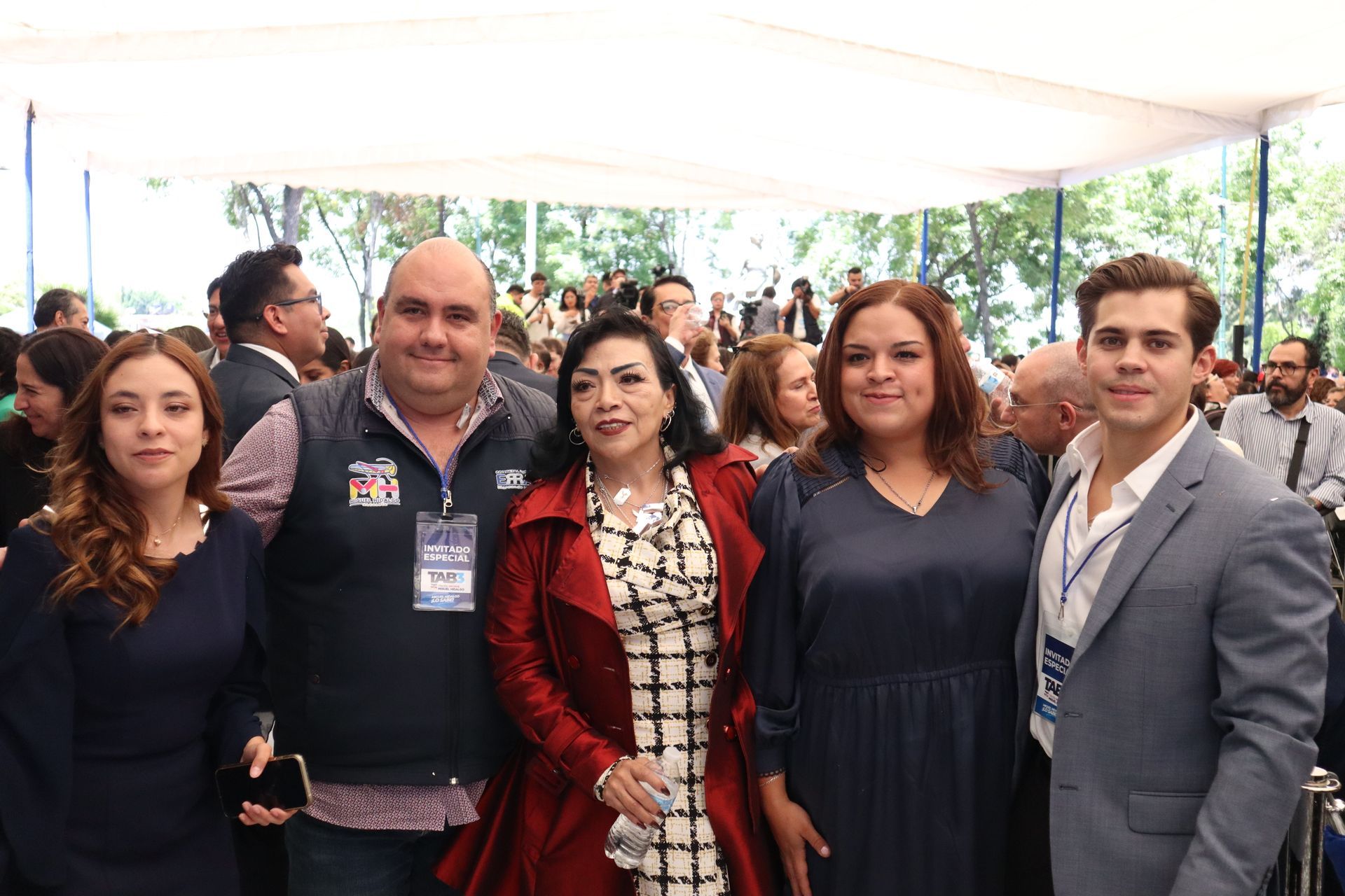 Five people posing together under a canopy at a crowded outdoor event, smiling at the camera.