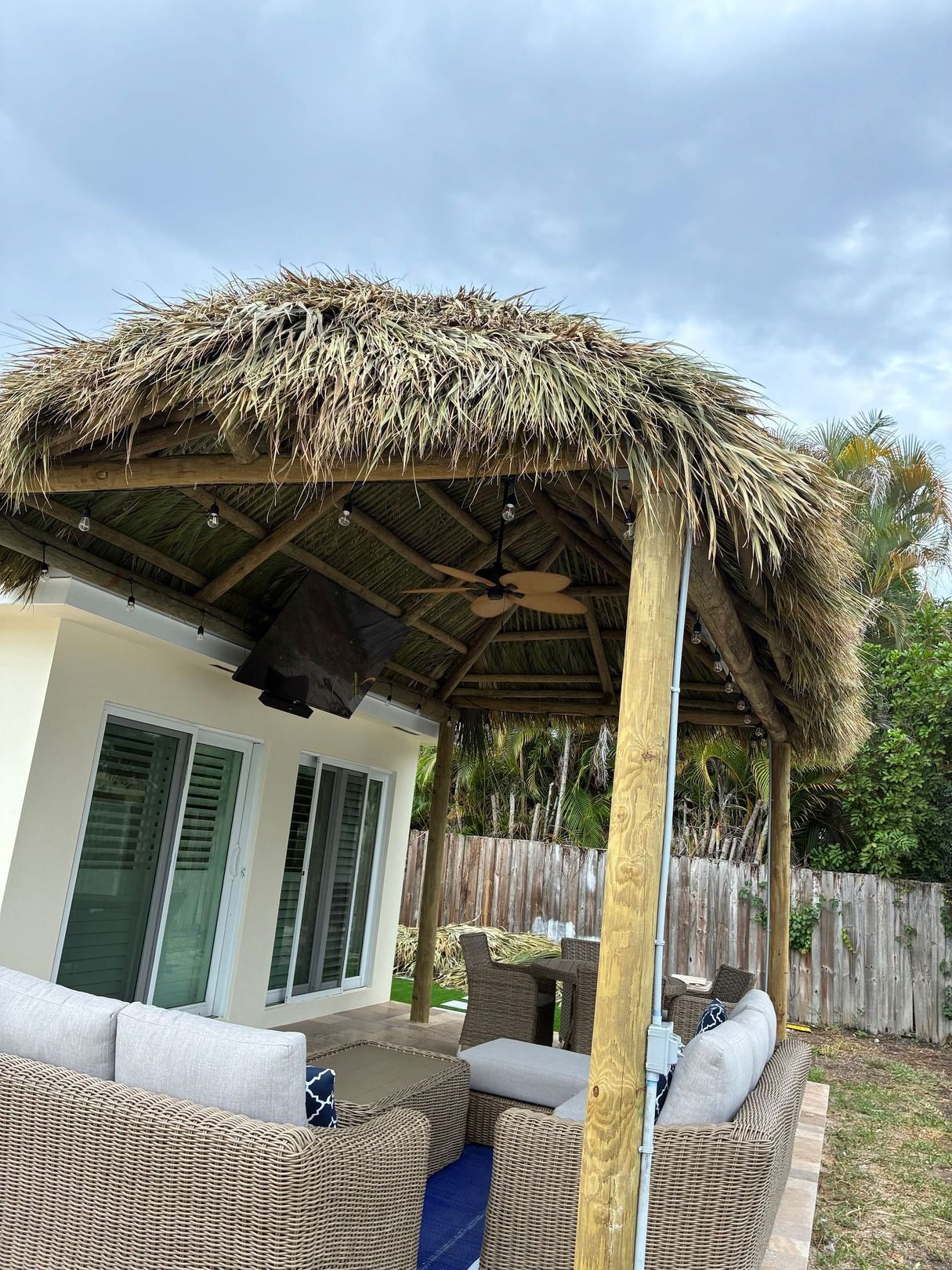 Tropical patio with thatched roof, wicker sofa, and white house with green shutters under a cloudy sky